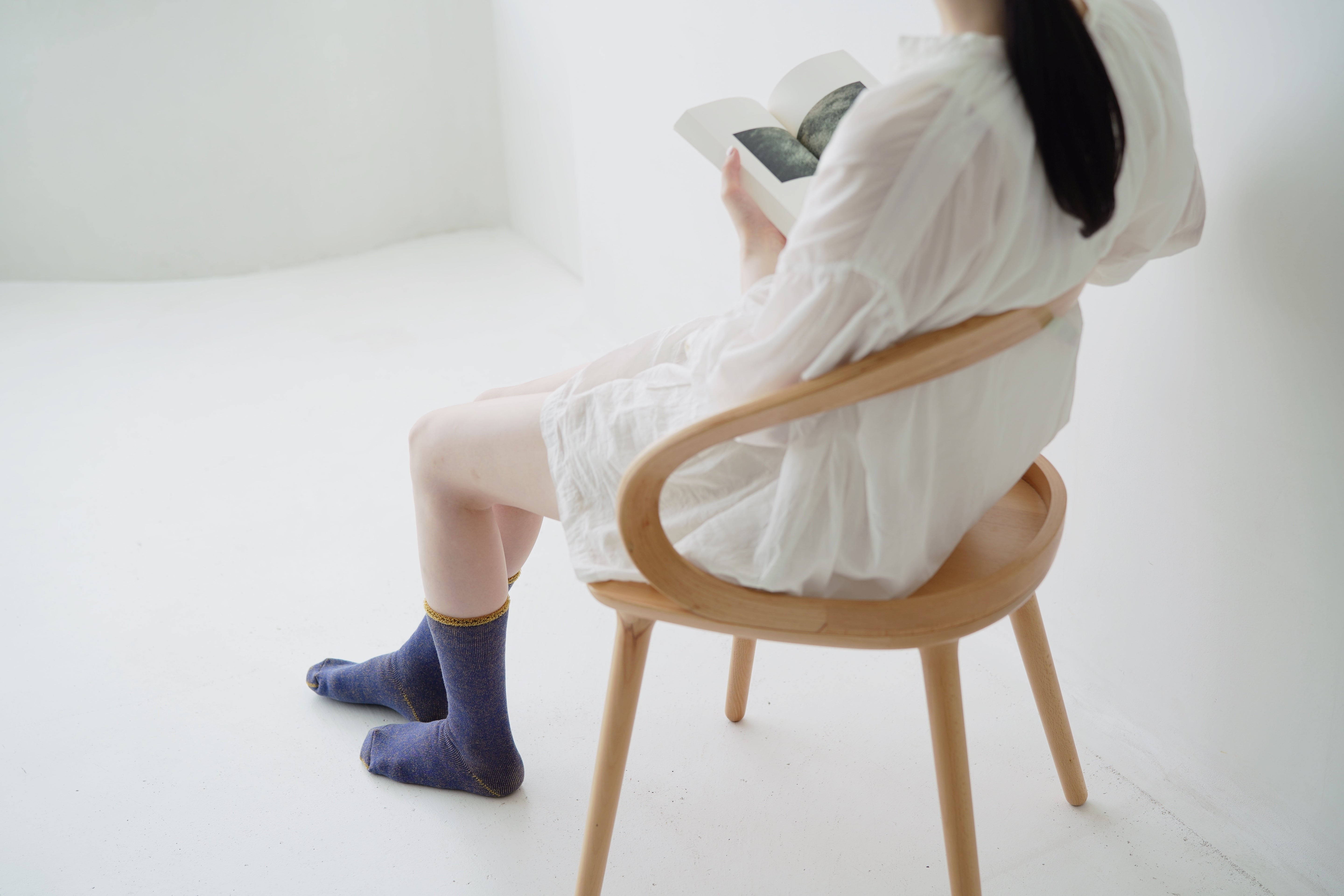 Woman in white dress seated in light wood chair, reading a book; wearing blue and gold socks.