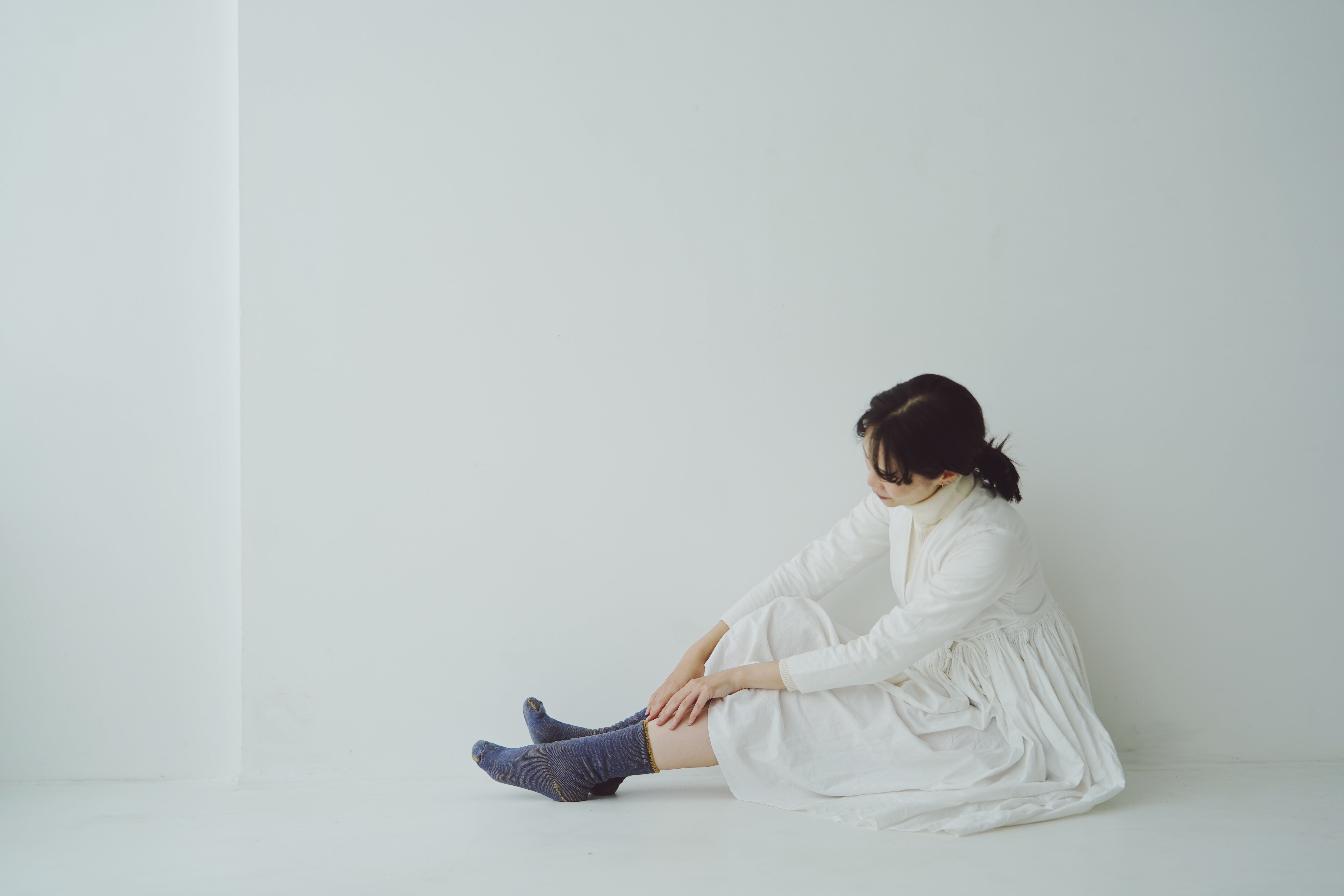 Woman in a long white dress sits on a white floor wearing blue socks with gold accents.