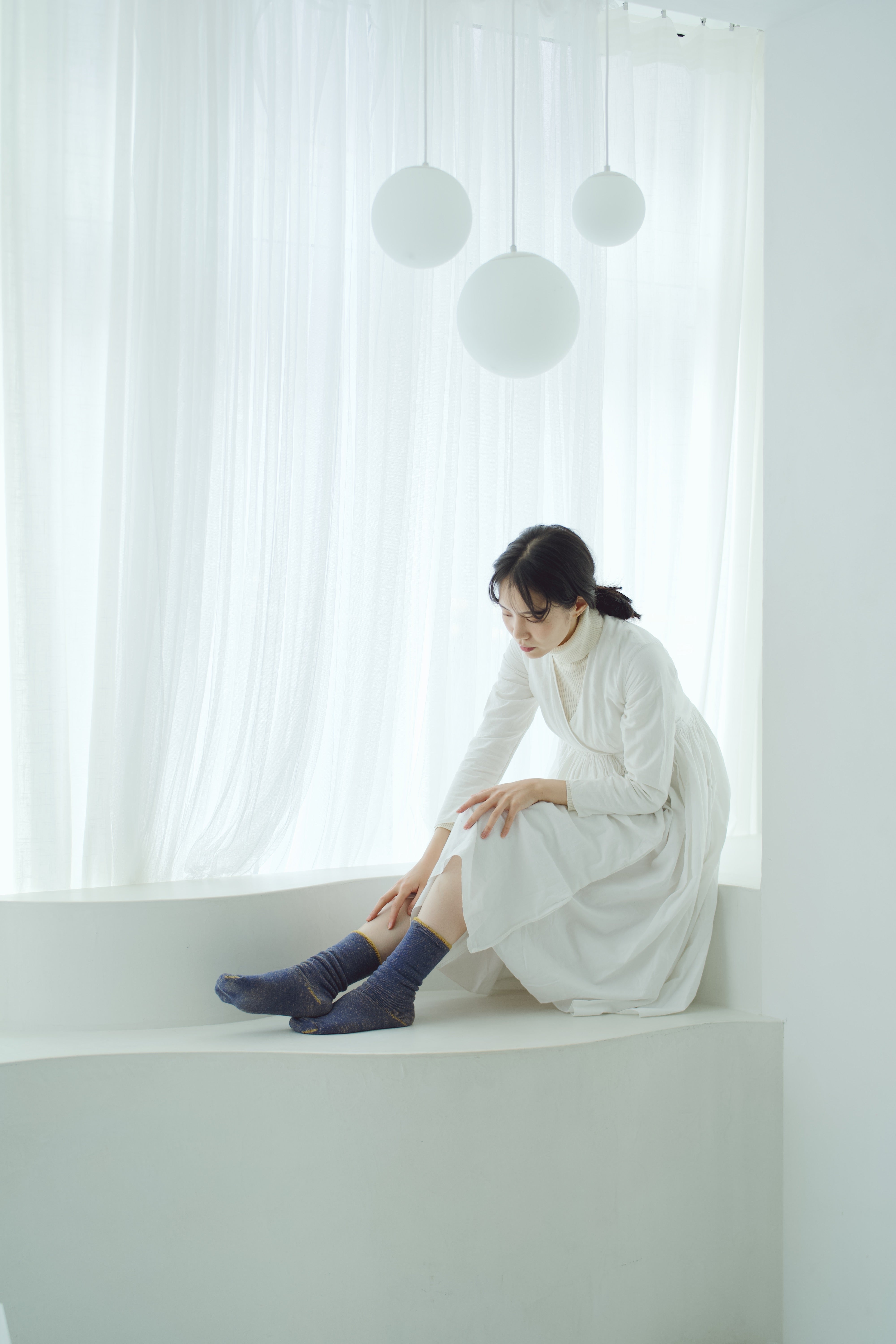 Woman in a white dress sits on a white window seat, wearing dark blue socks. Three round pendant lights hang above.