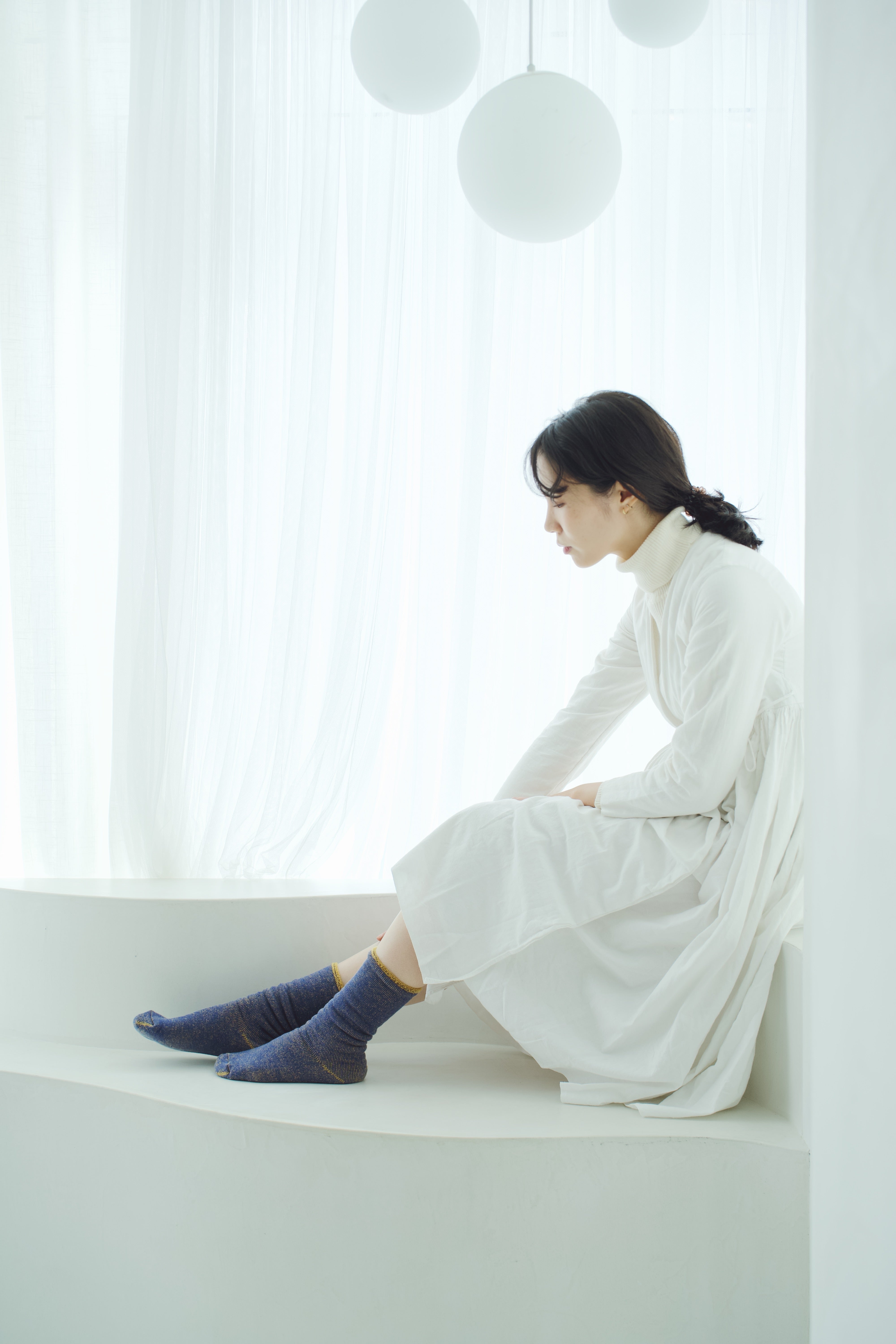 Woman in a white dress and blue socks sits on a white window seat.