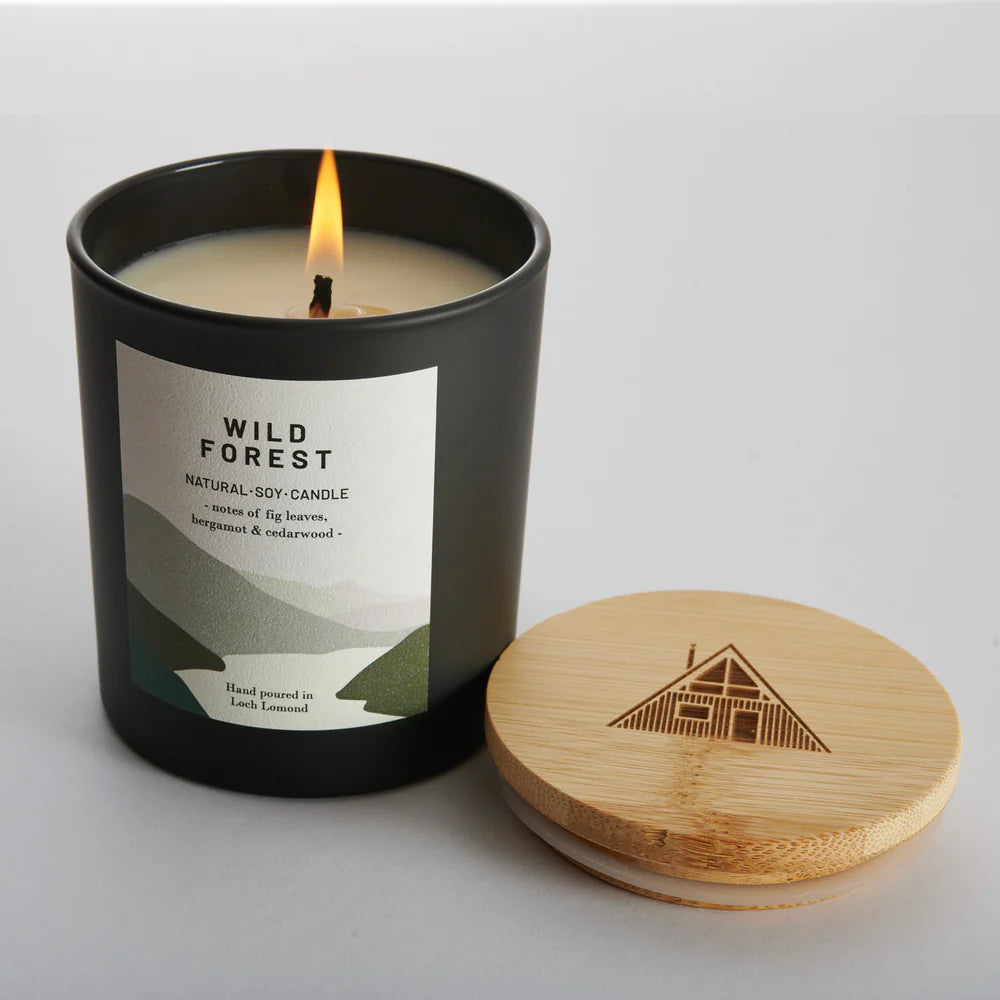 Black matte jar candle, lit, labeled "Wild Forest," natural soy blend with fig, bergamot & cedarwood scents.