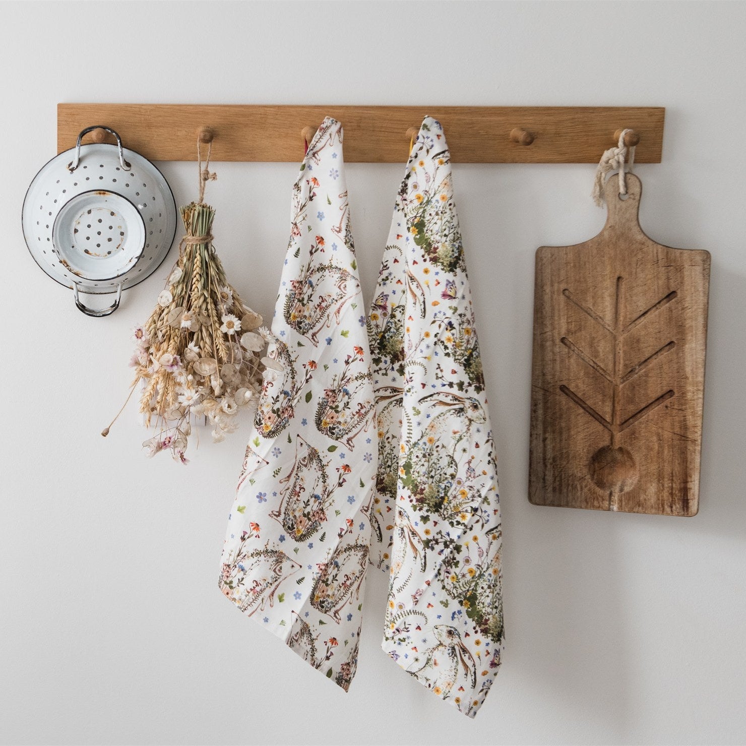 Pair of white tea towels with floral and hare print, hanging on a wooden rack.