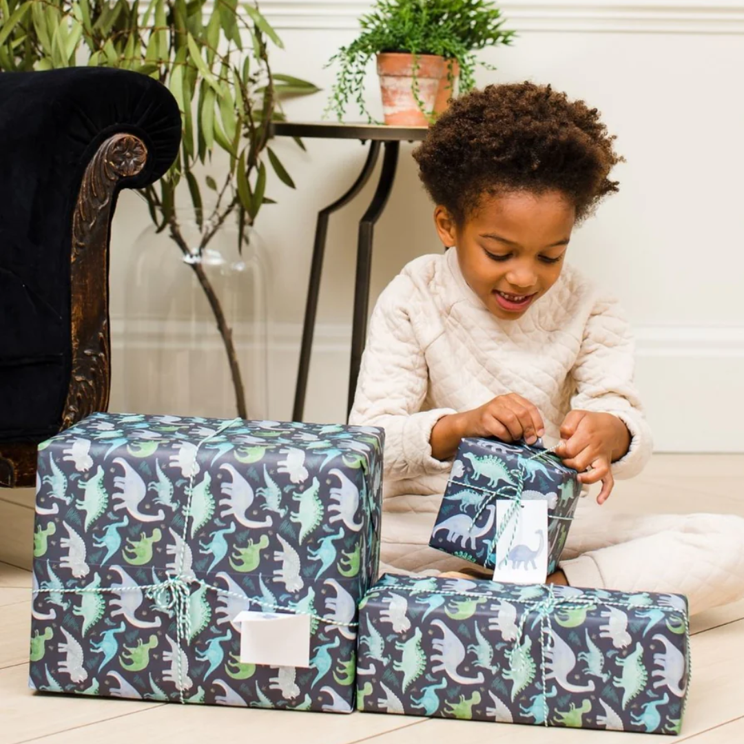 Three gifts wrapped in dark-blue dinosaur-patterned paper; a child is opening one.