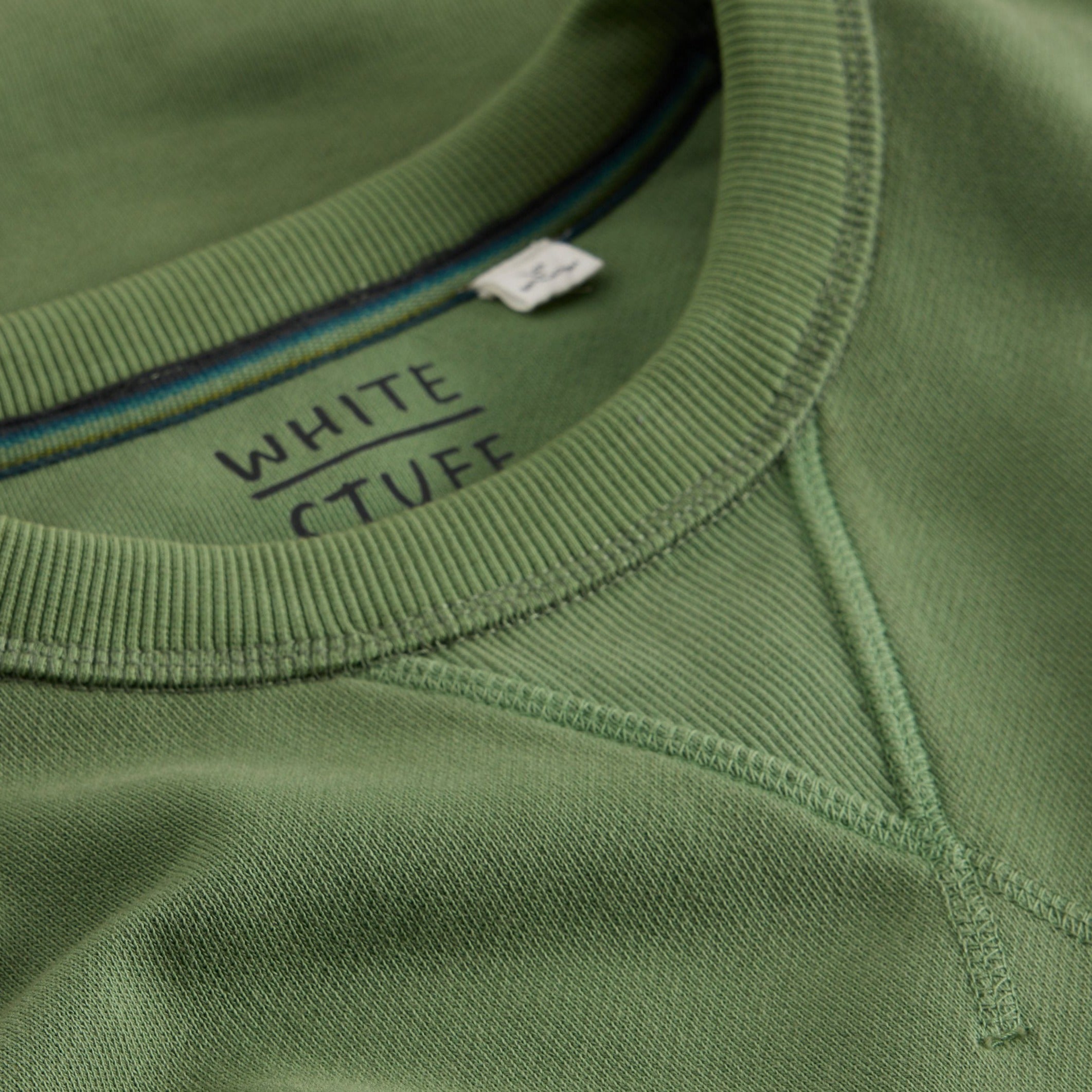 Close-up of a sage green crewneck sweatshirt's neckline and shoulder seam, showing ribbed fabric and "WHITE STUFF" label.