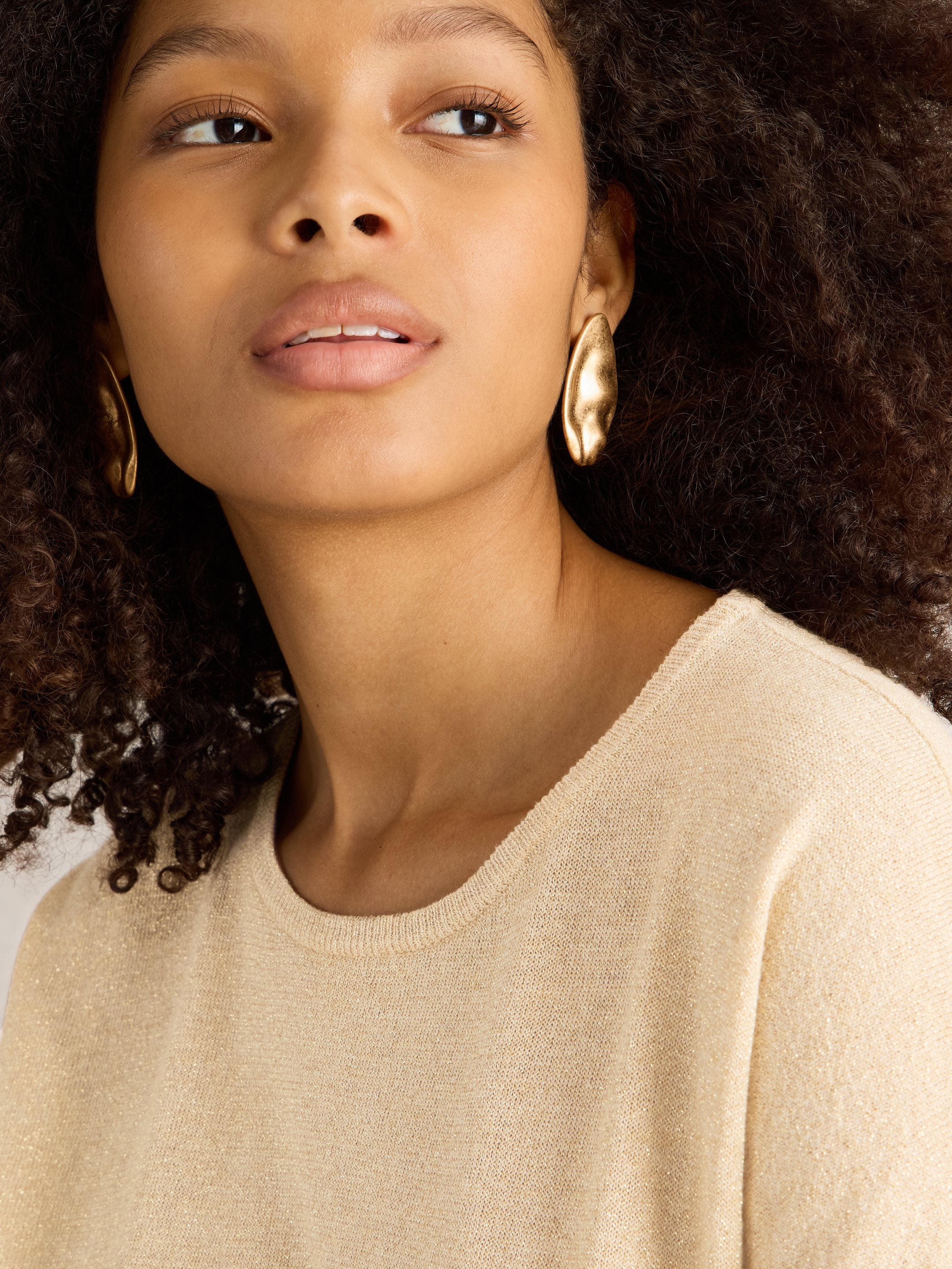 Beige sparkly knit sweater with gold, abstract-shaped earrings.