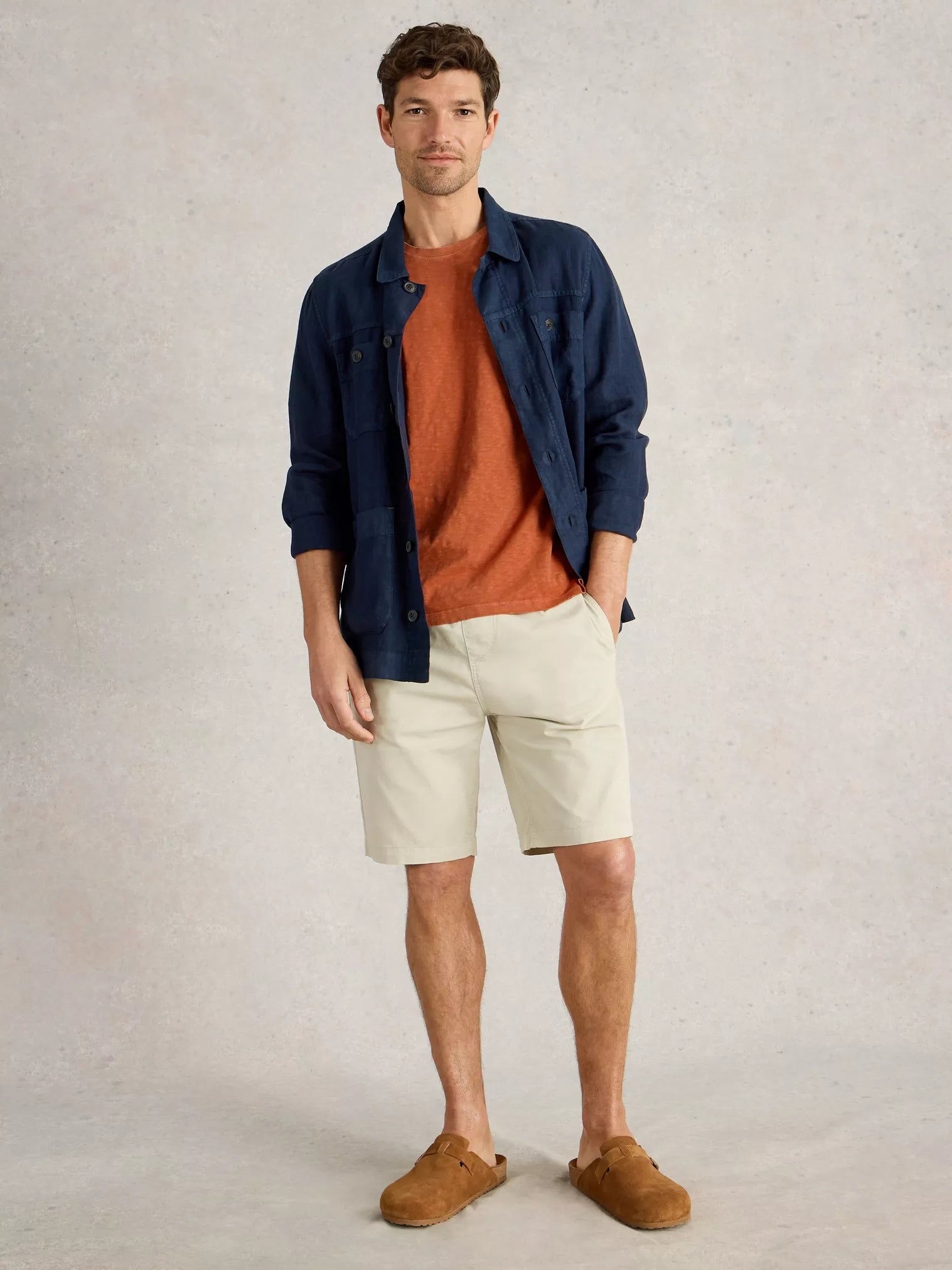 Man wearing beige chino shorts, a rust-colored t-shirt, and a navy blue linen shirt jacket. He's wearing brown suede clogs.