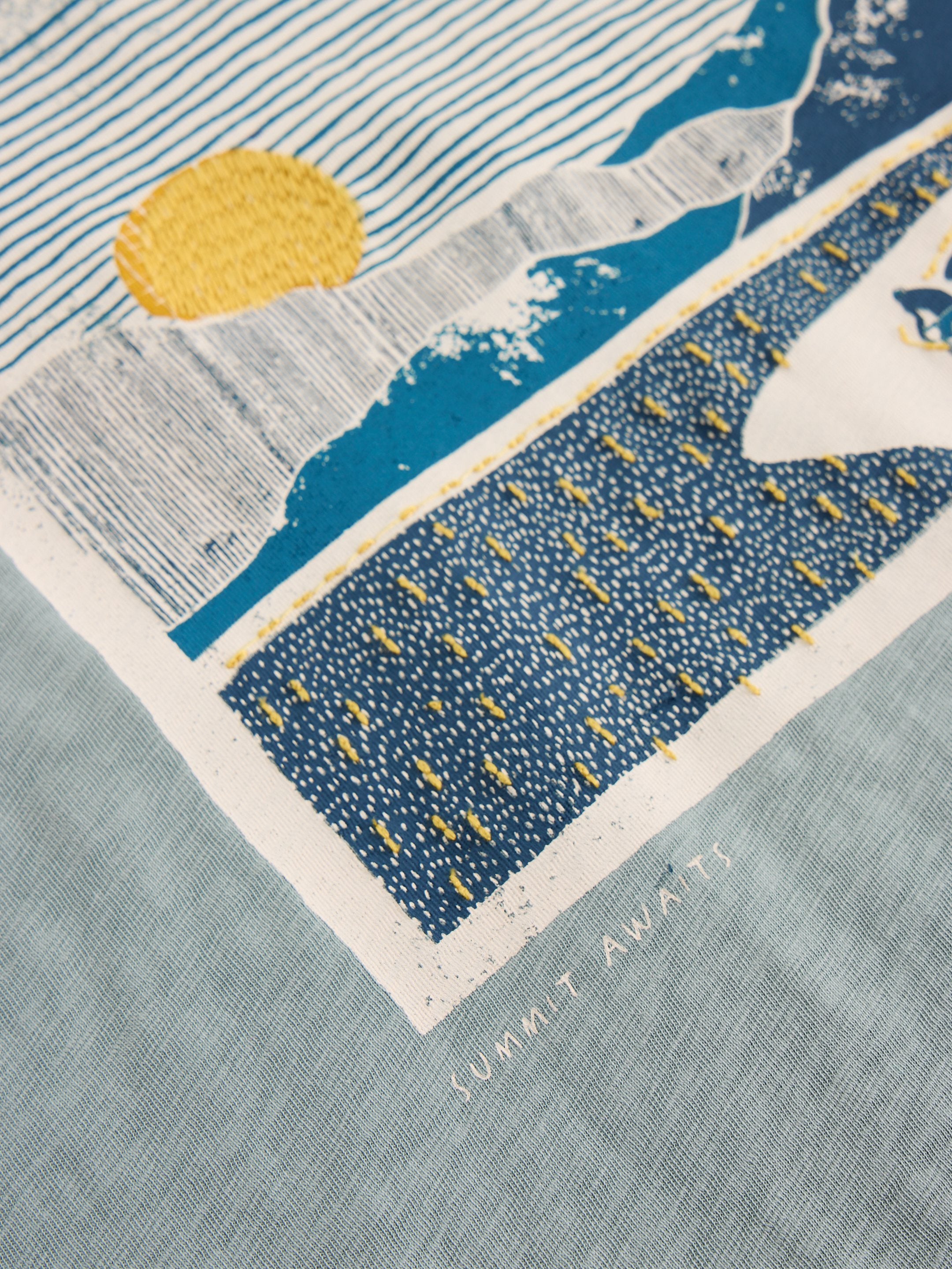 Close-up of a teal shirt featuring an embroidered and printed landscape design; a yellow sun, blue mountains, and textured