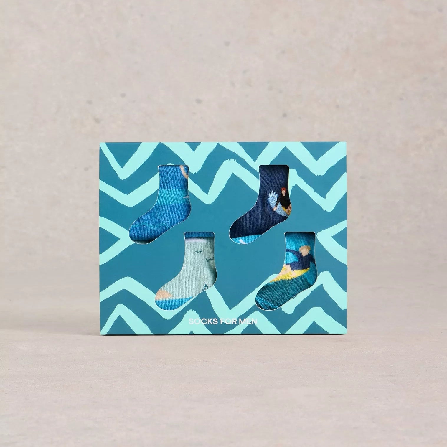 Four pairs of men's socks in a teal gift box with a zig-zag pattern. Each sock features a different ocean-themed design.