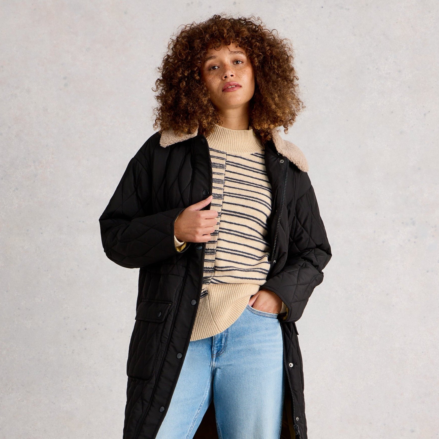 Woman wearing a black coat, striped shirt, and blue jeans on a plain background