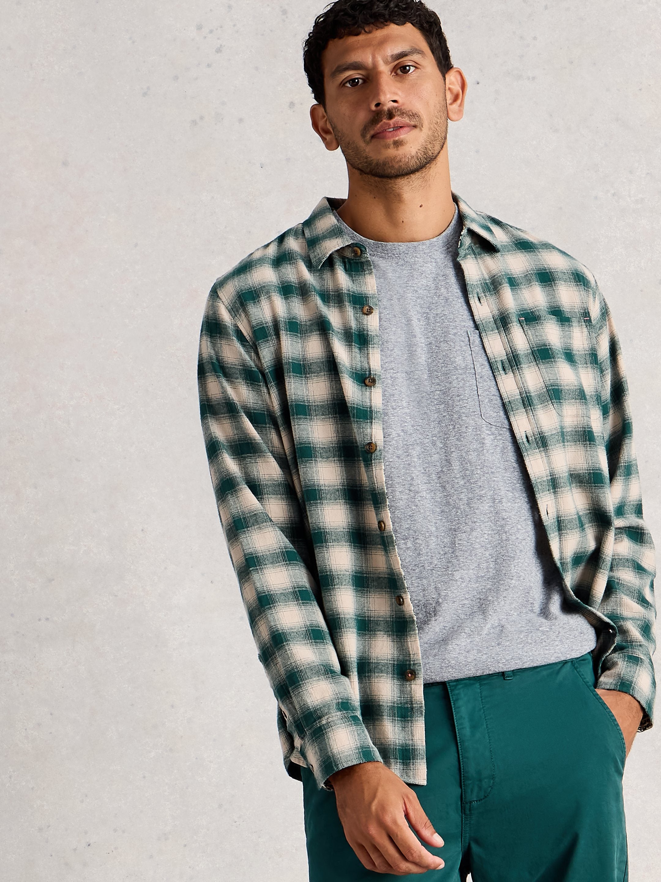 Man wearing a plaid shirt over a gray sweater with teal pants against a light gray background