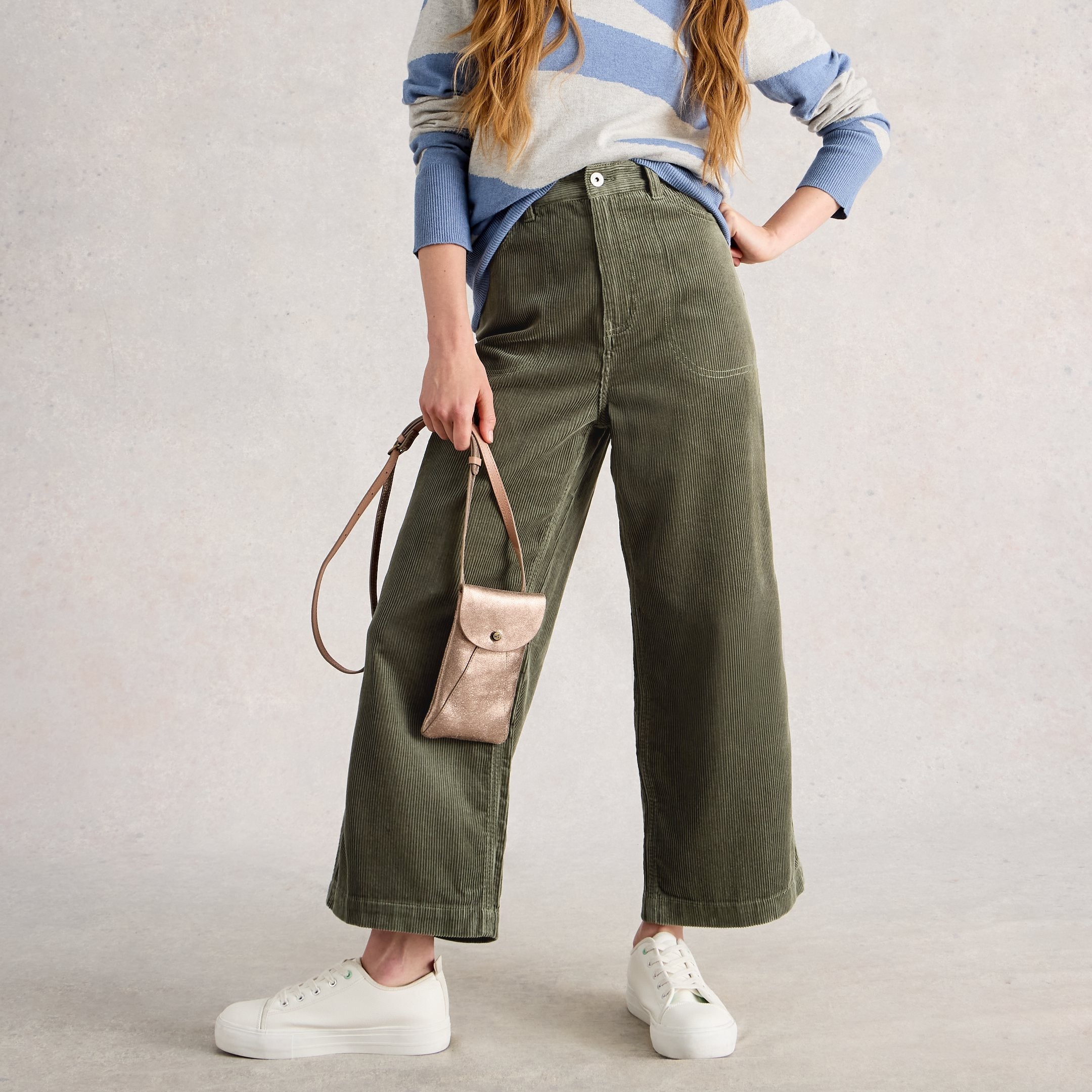 Woman wearing a striped sweater and green pants with a beige handbag on a light background