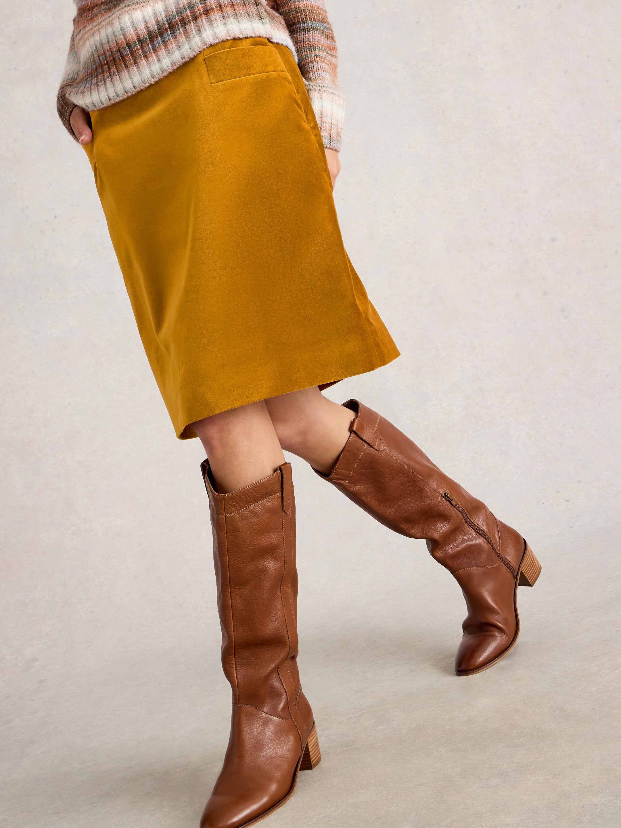 Person wearing mustard skirt and brown knee-high boots on a neutral background