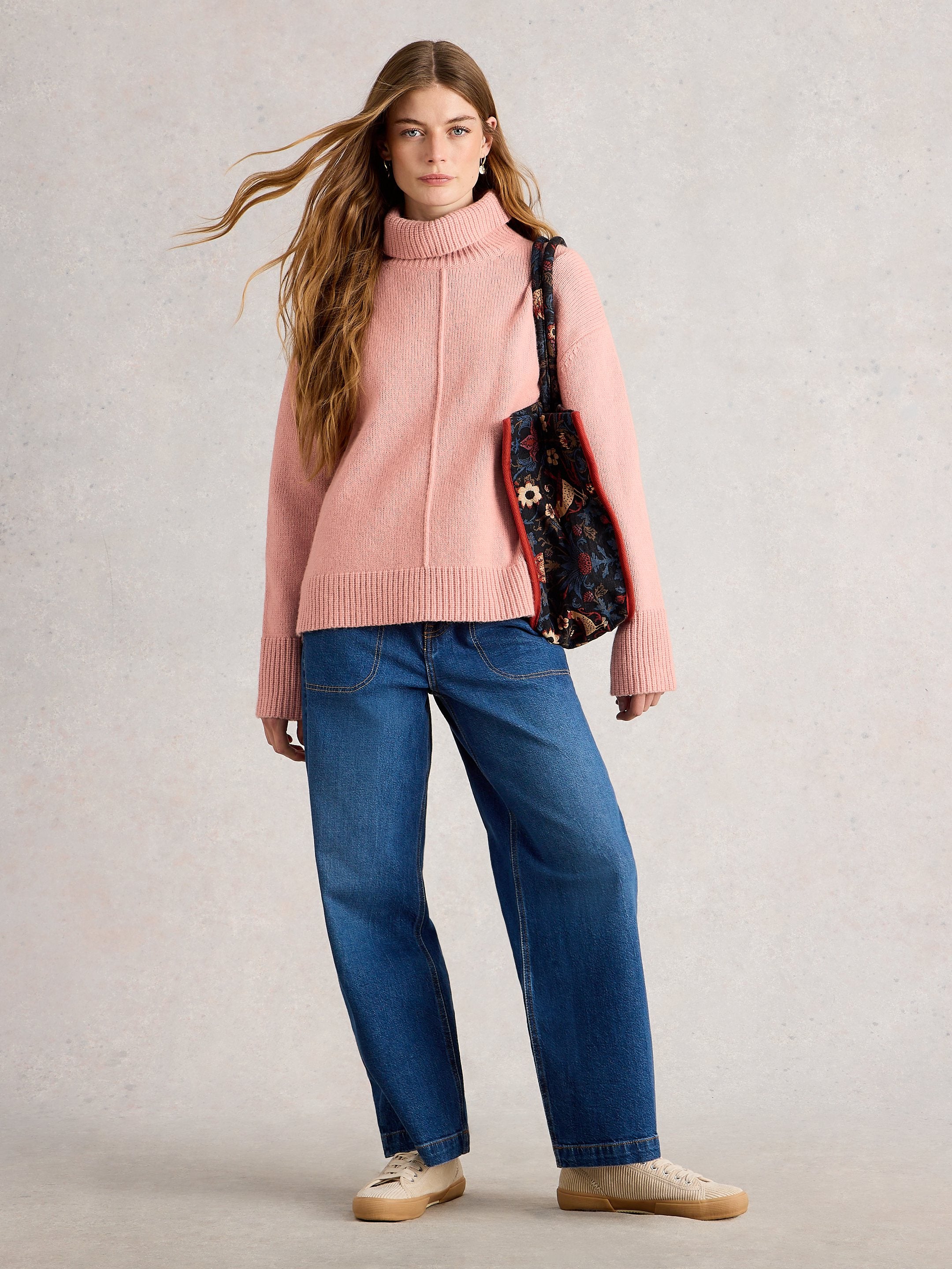 Woman wearing a pink sweater and blue jeans with a patterned bag on a light gray background