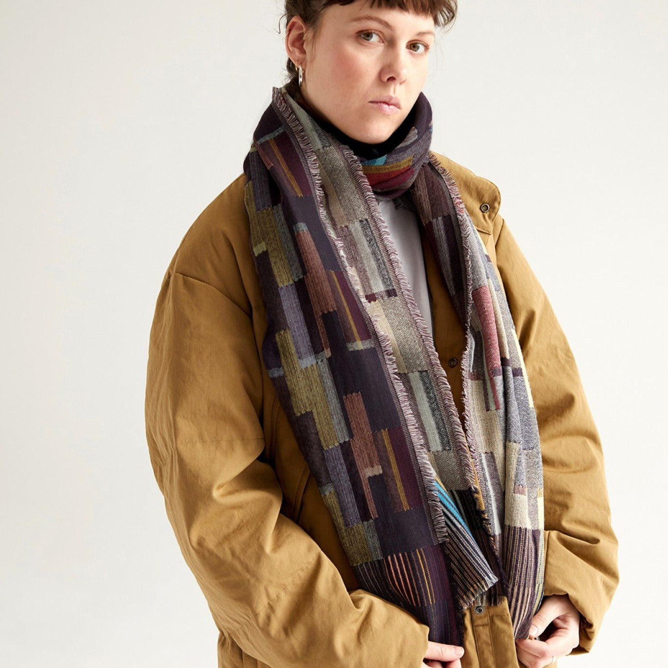 Woven scarf with a geometric pattern in shades of purple, gray, and beige.  The scarf has a fringed edge.