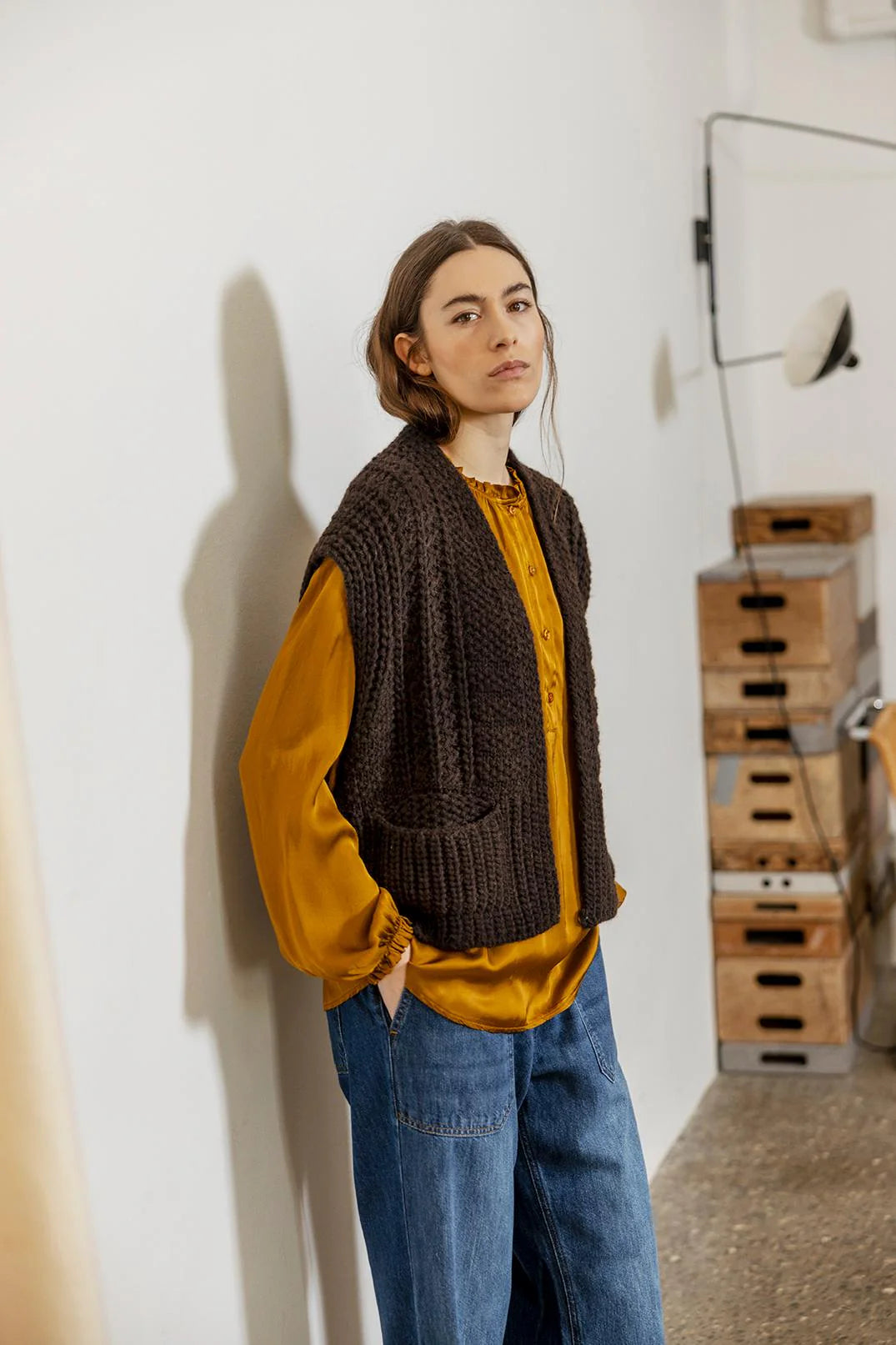 Person wearing a brown knitted vest over a mustard shirt with blue jeans, standing against a white wall with wooden crates in the background.