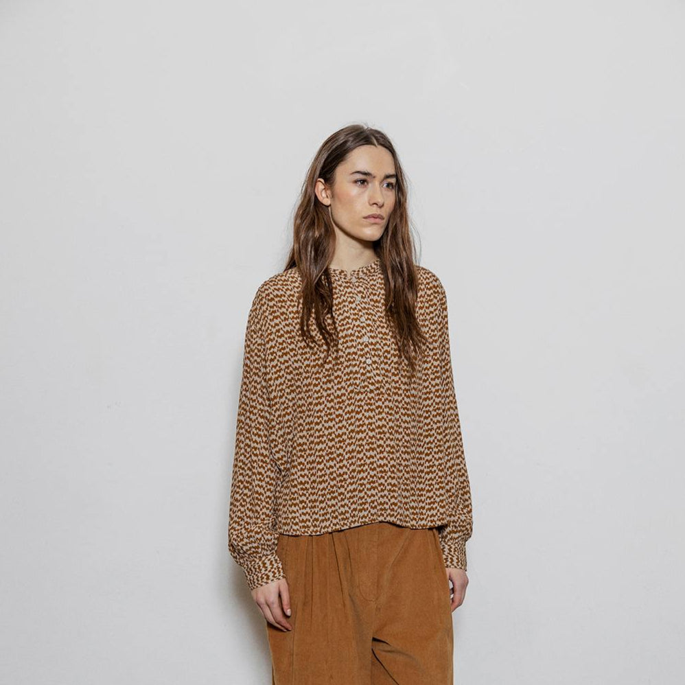 Person wearing a patterned brown sweater and brown pants against a white wall.