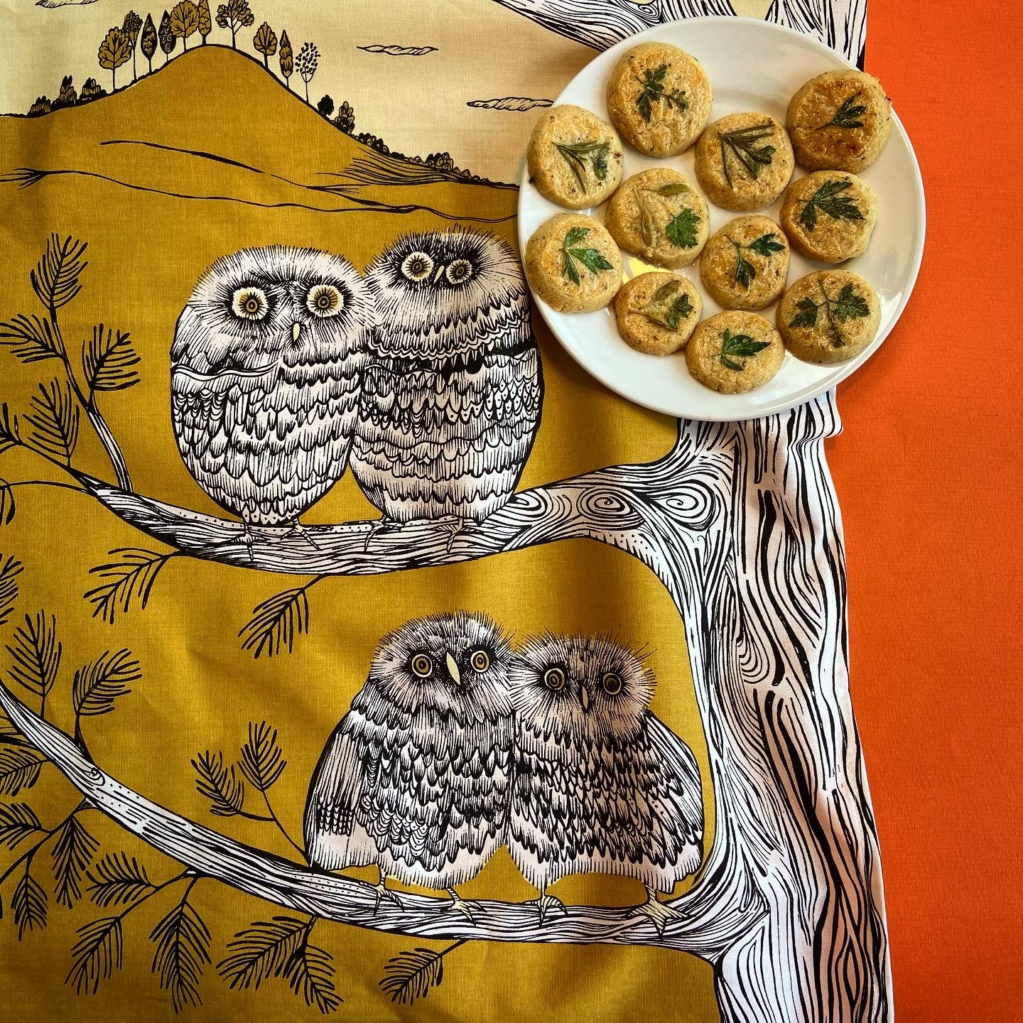 A plate of round, herb-topped crackers sits atop a mustard-yellow fabric featuring a whimsical owl design.