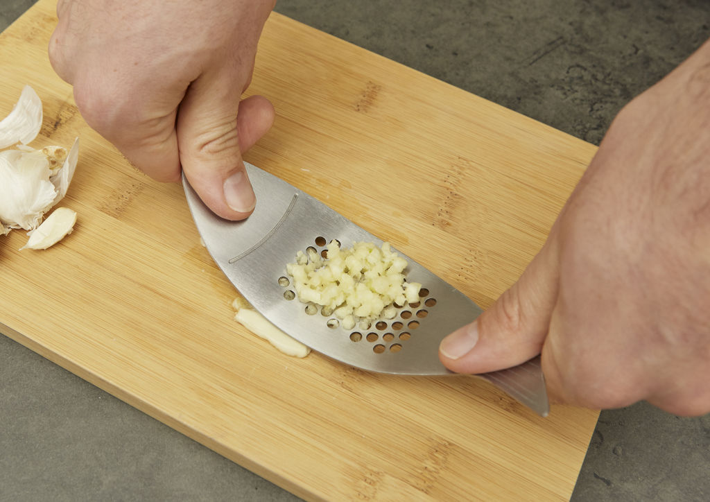 Stainless steel garlic press with minced garlic on a bamboo cutting board.