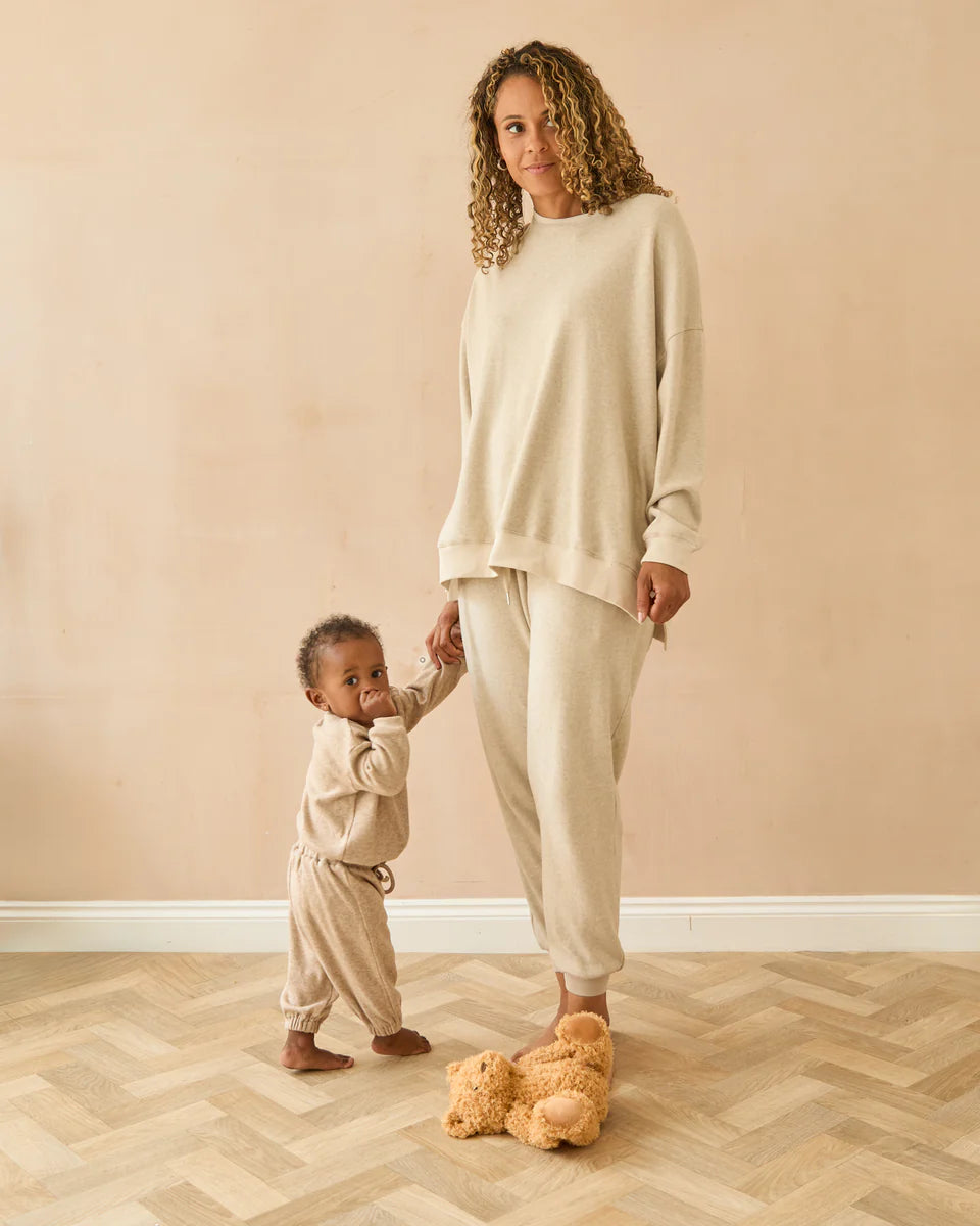 Woman and child in matching beige outfits standing on a wooden floor against a beige wall.