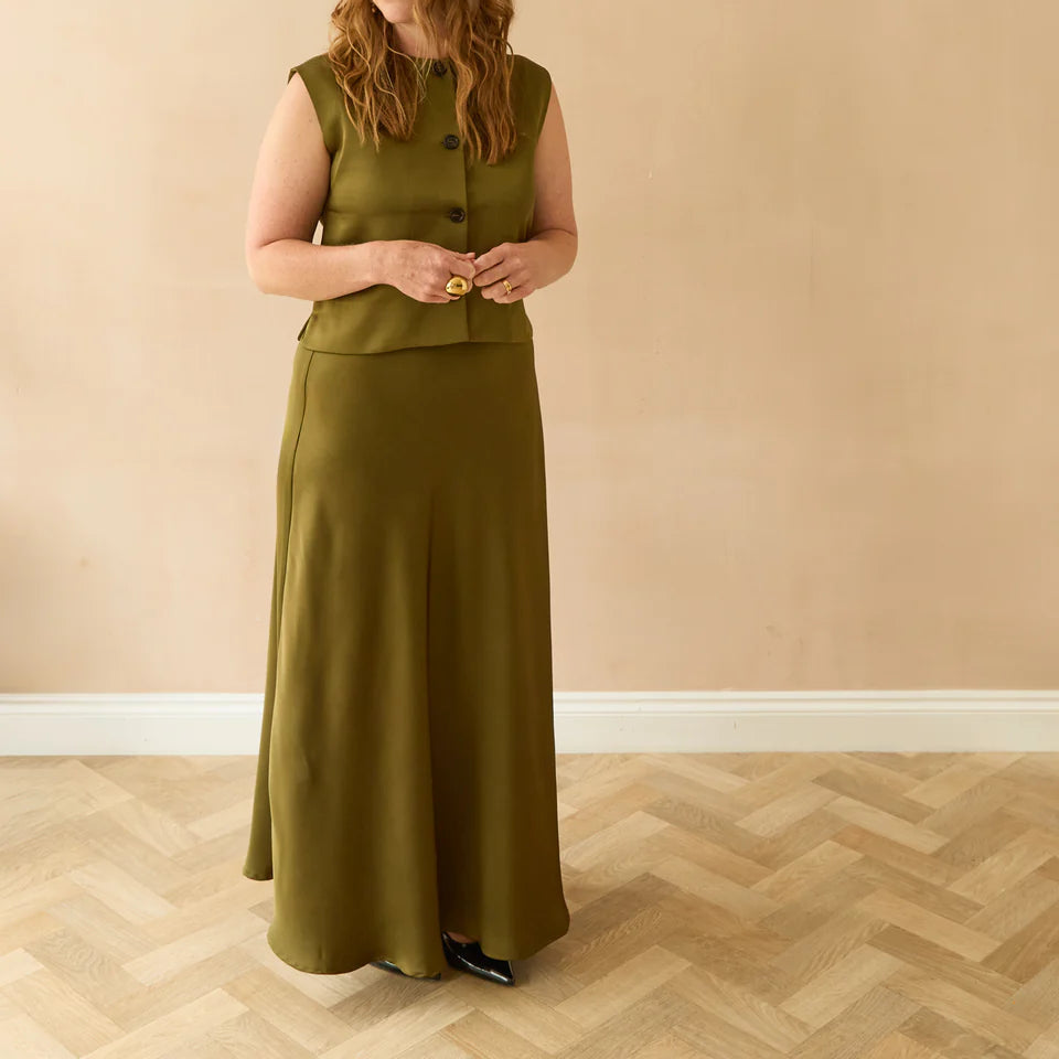 Woman wearing a green sleeveless top and long skirt standing against a beige wall.