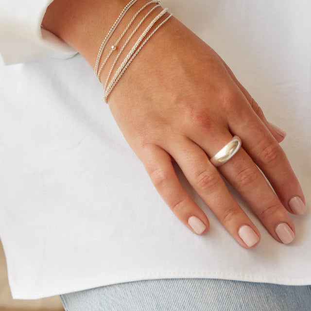 Silver ring with a smooth, rounded, dome-shaped band, worn on a finger alongside several delicate silver chain bracelets.