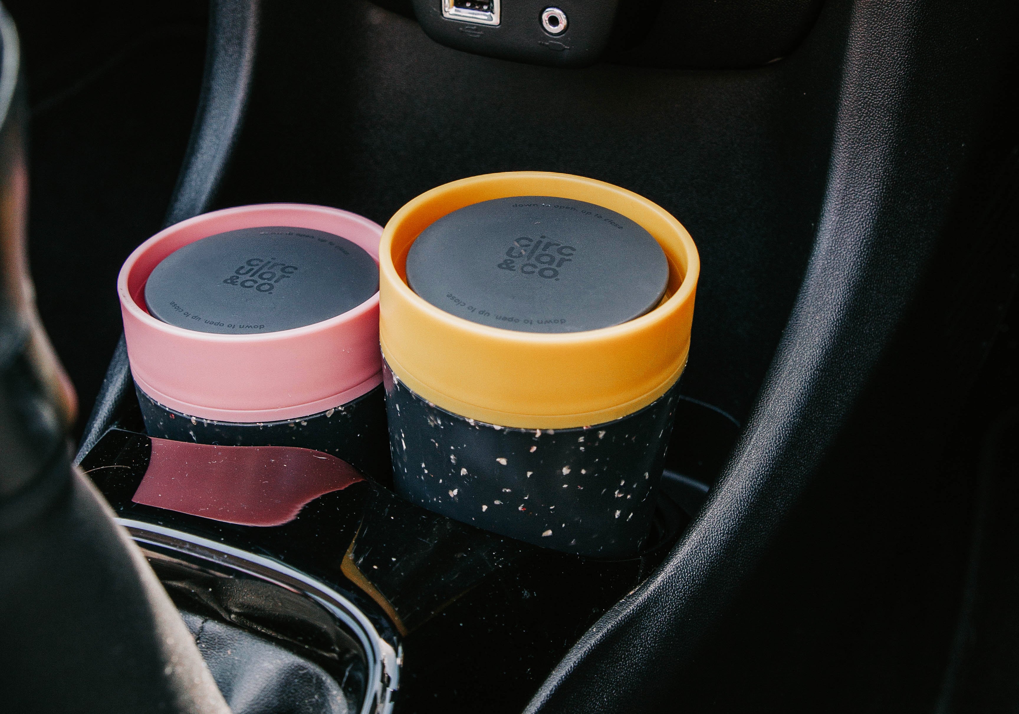 Two Circulon & Co. reusable coffee cups with speckled black bodies, pink and mustard lids, in a car cup holder.