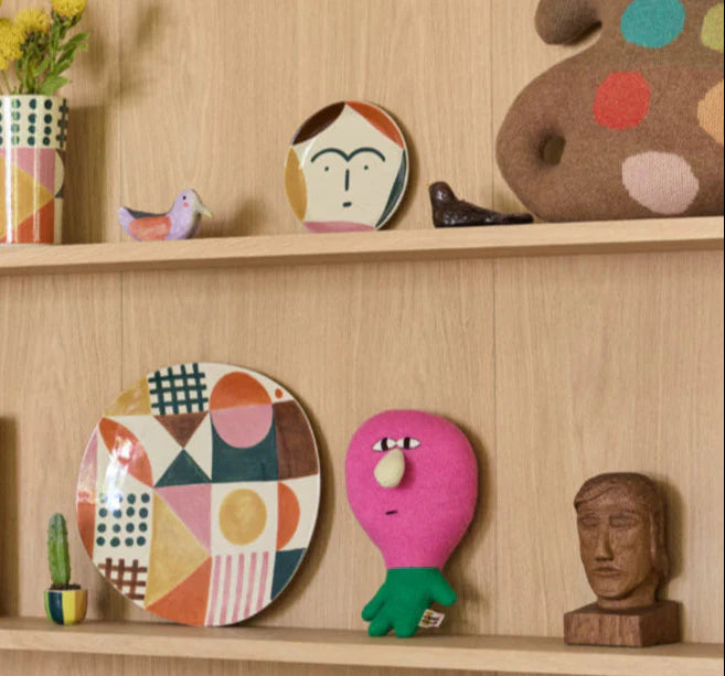 Decorative shelves with colorful plates, a pink plush toy, and wooden figurines on a wooden shelf.
