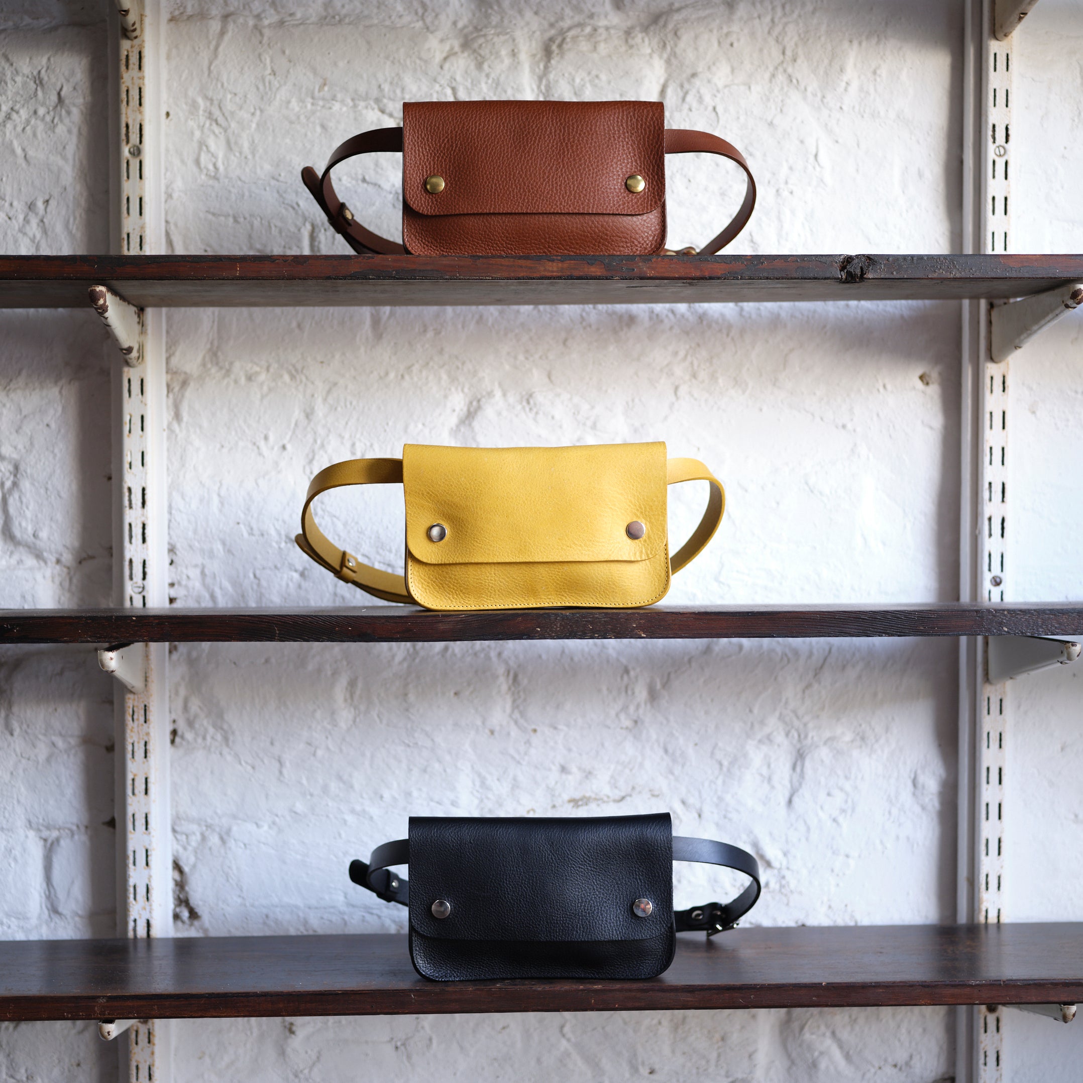 Three leather fanny packs in brown, yellow, and black displayed on wooden shelves.