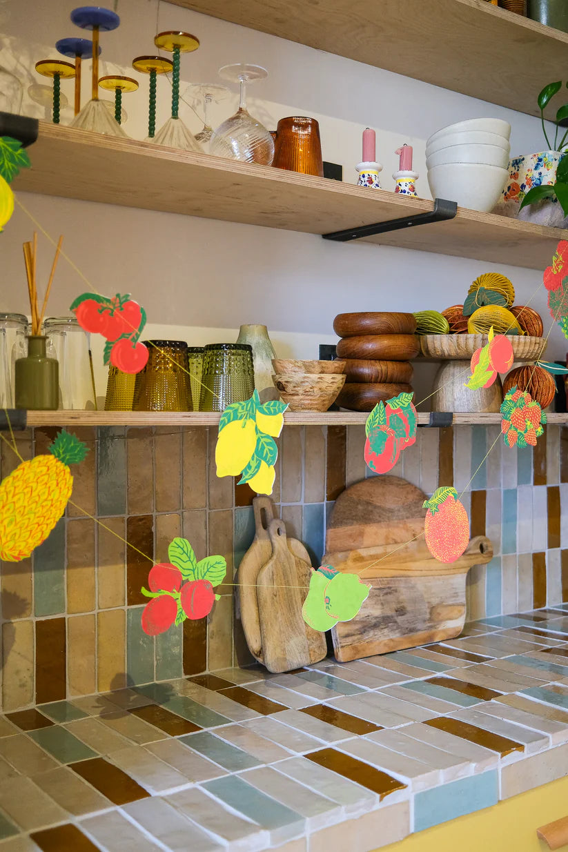 Fruit-themed paper garland strung below kitchen shelves stocked with glassware, wooden bowls, and other kitchenware.
