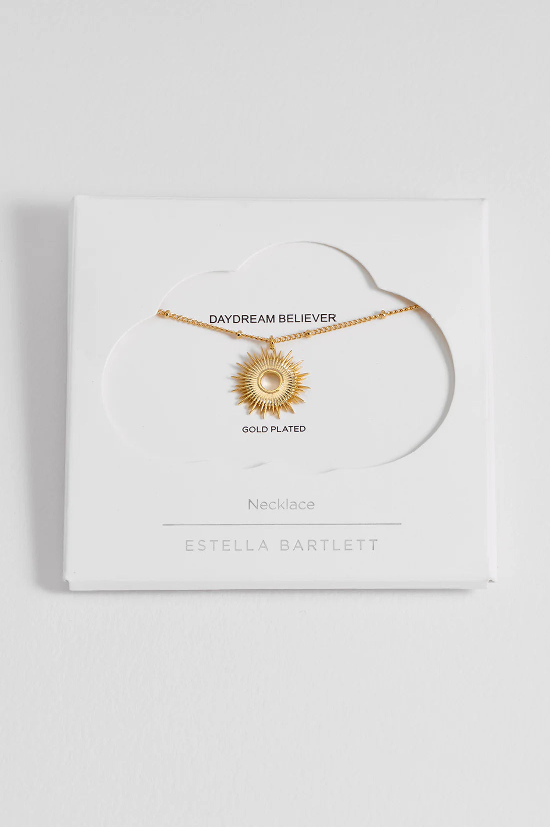 Gold-plated sunburst necklace on white card packaging.  "Daydream Believer" and "Estella Bartlett" branding visible.