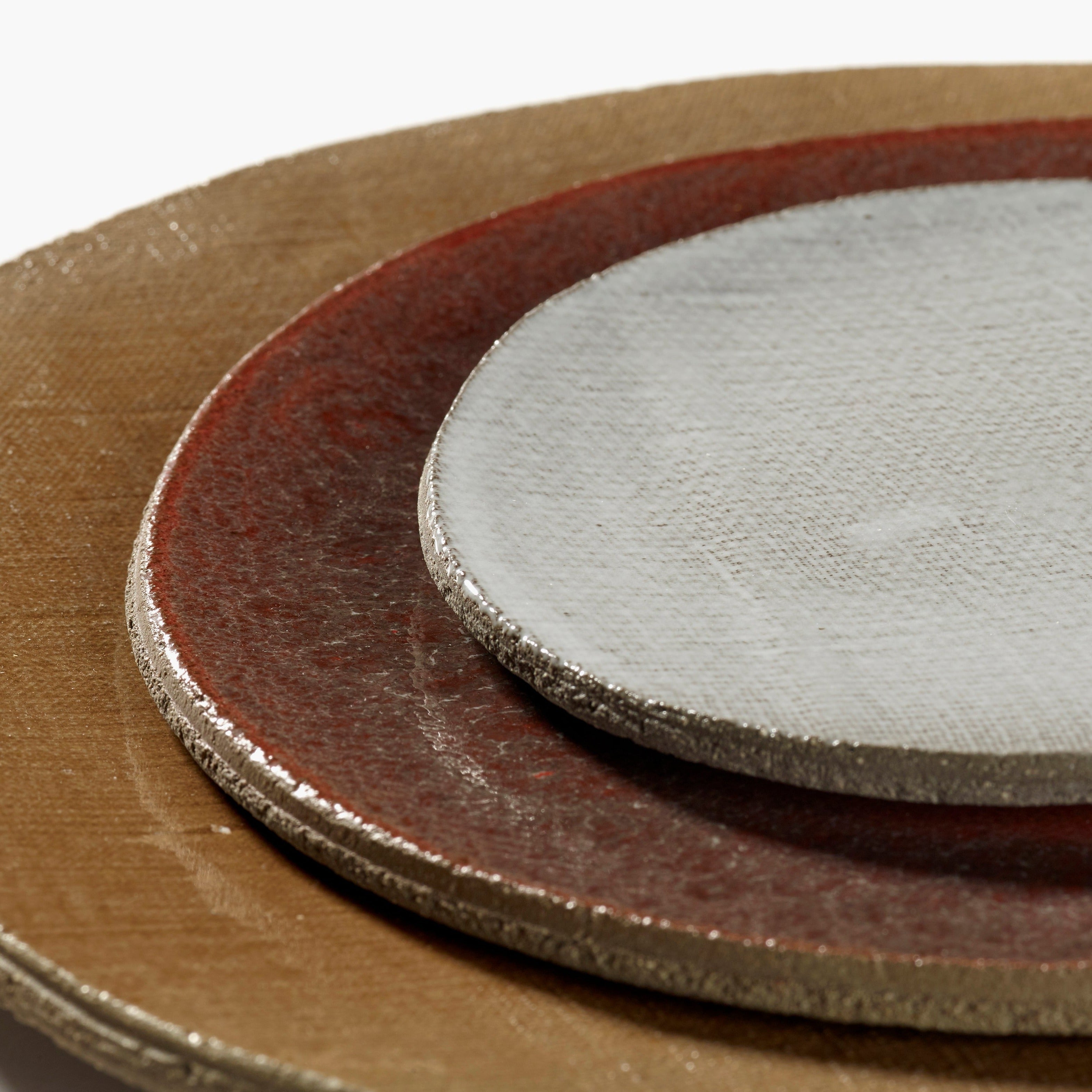 Three ceramic plates stacked, in off-white, reddish-brown, and tan.  Each plate has a textured, slightly irregular edge.