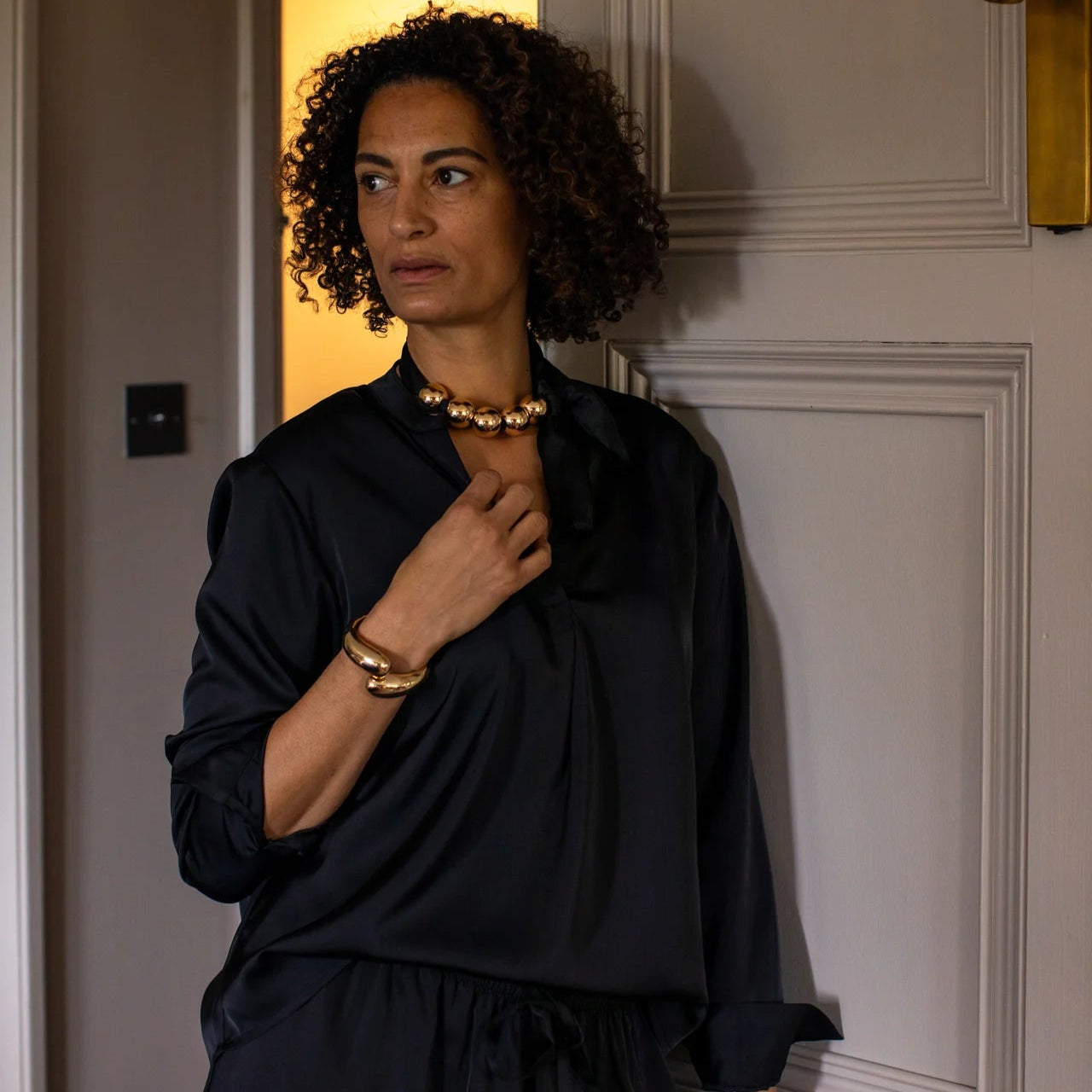 Woman wearing a black outfit with gold jewelry against a neutral wall.
