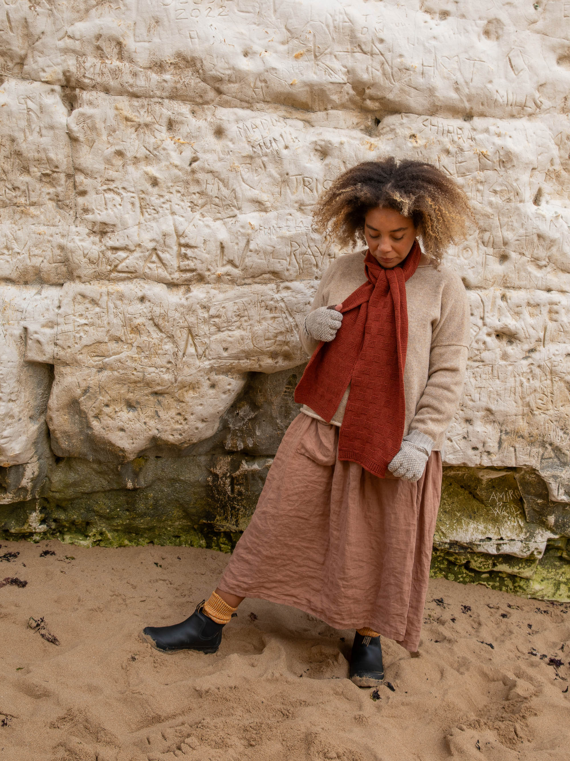 A woman stands on a sandy beach wearing a rust-colored knitted scarf, a long mauve skirt, and black ankle boots.