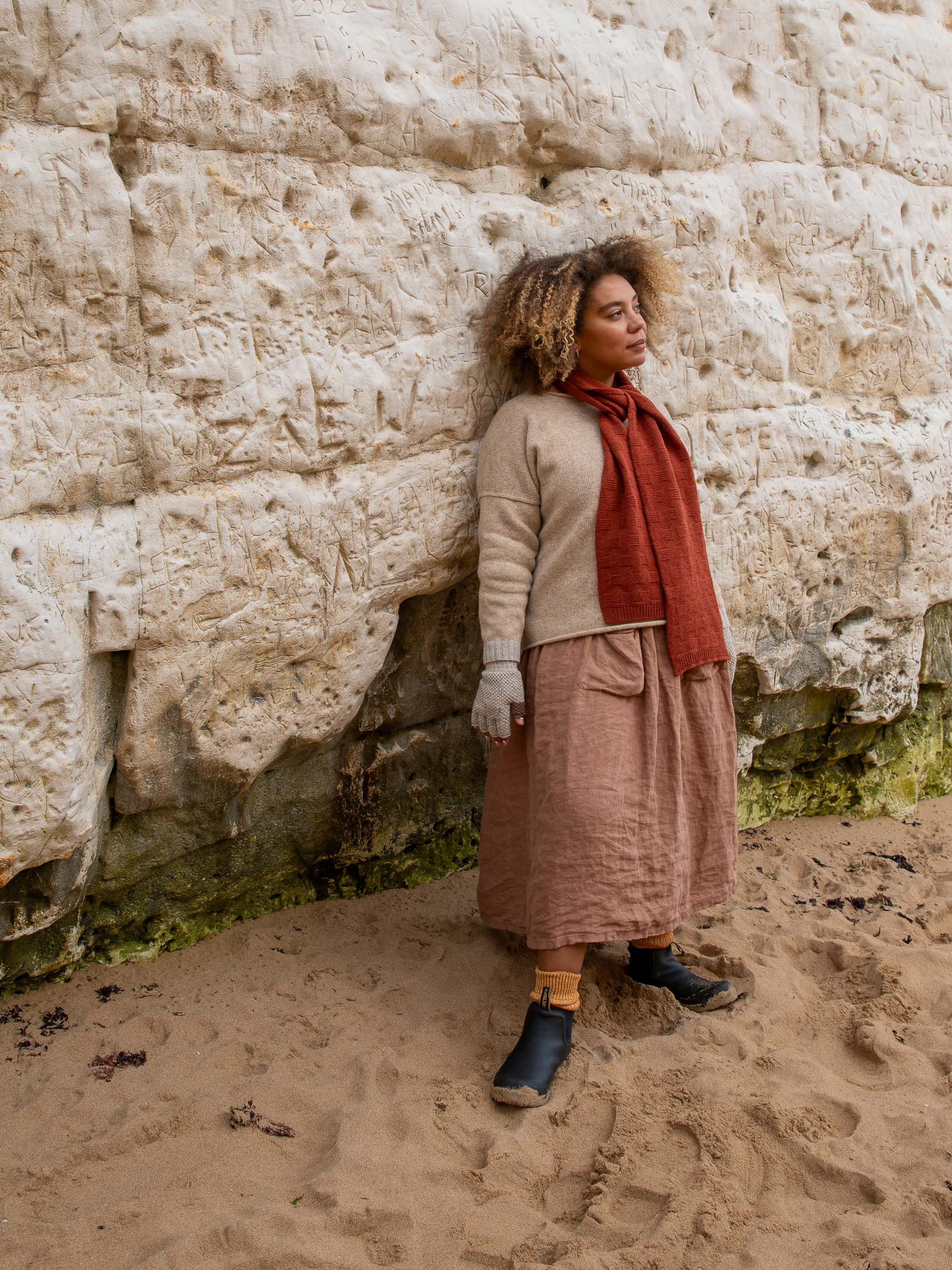 Woman wearing a beige sweater, rust-colored scarf, mauve linen midi skirt, and black ankle boots stands against a textured,