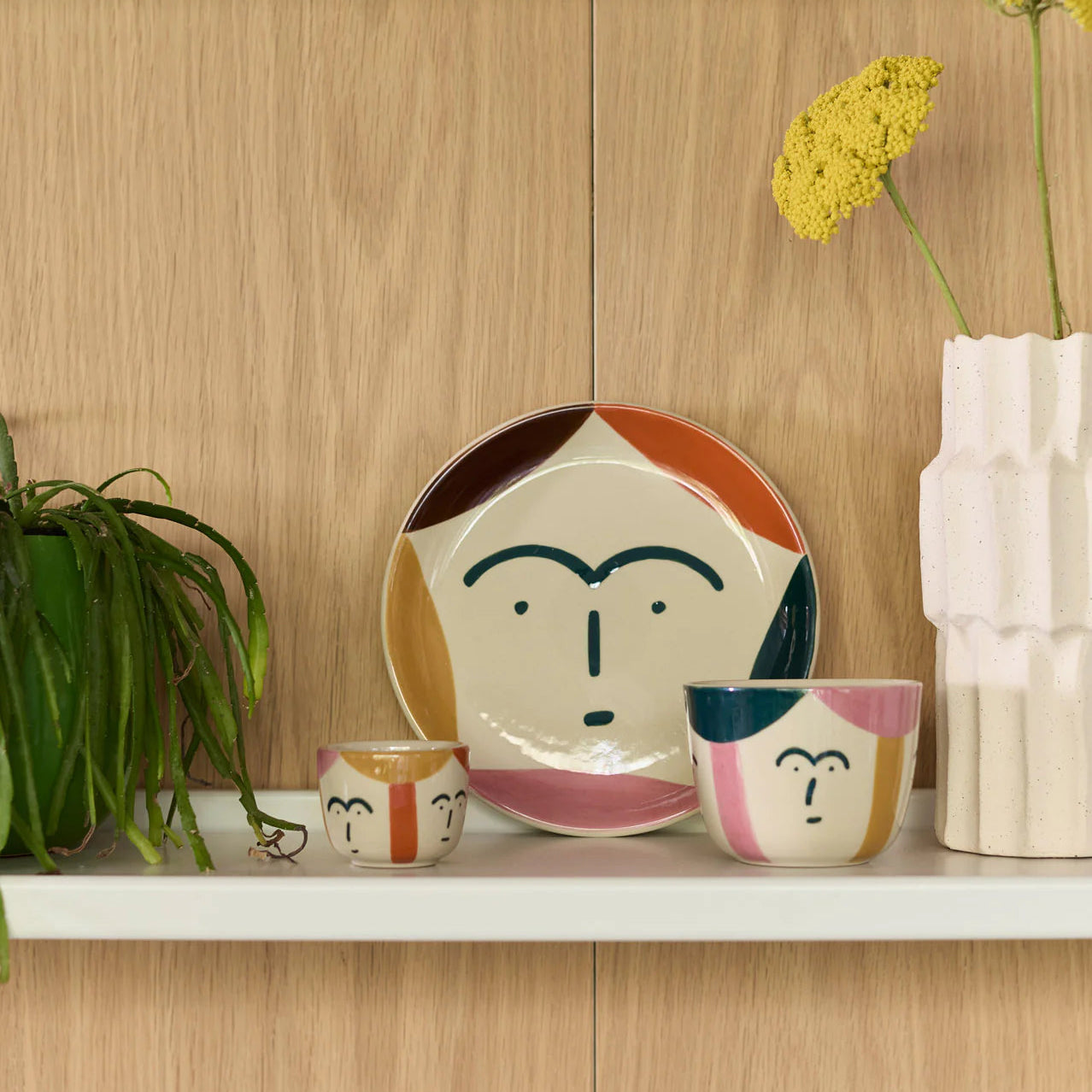 Ceramic face-themed plates and bowls on a wooden shelf with a plant and vase in the background.