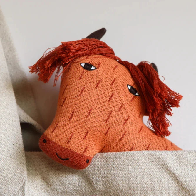 Orange plush horse head with red hair, designed by Donna Wilson, peeking out from a light beige fabric background.