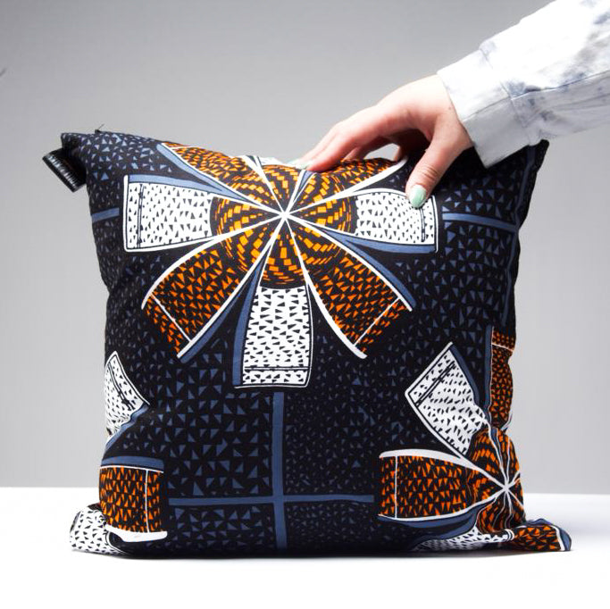Square decorative pillow with a black background and a repeating pattern of orange, white, and blue stylized flowers.
