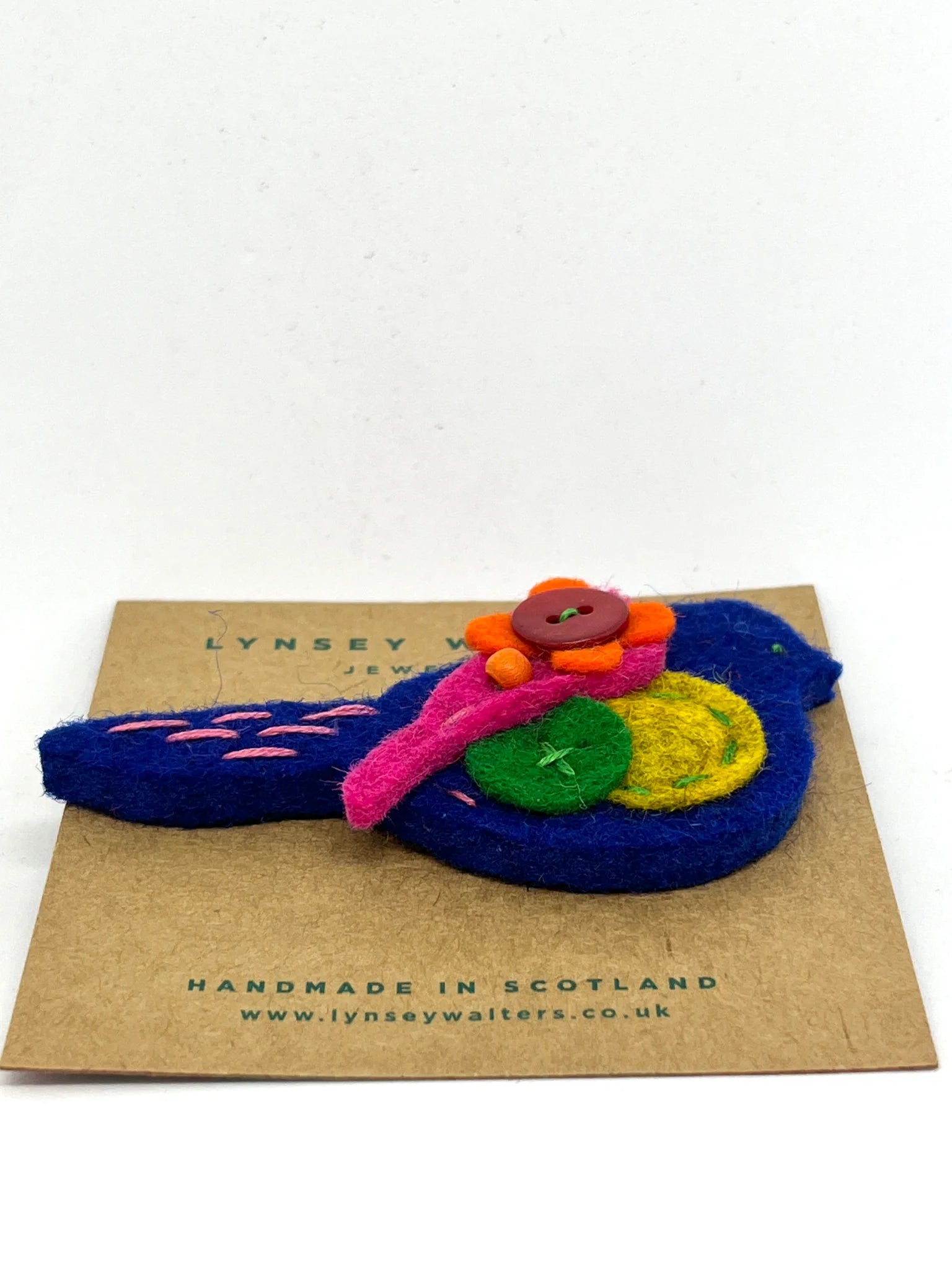 Handmade felt bird brooch in blue, adorned with pink, orange, yellow, and green felt flowers and buttons.