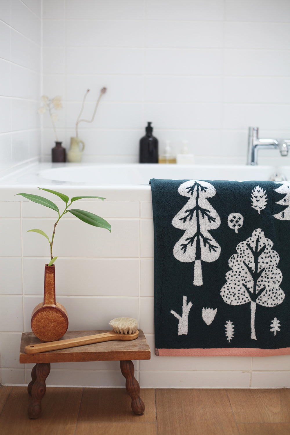 A dark green towel with white tree designs by Donna Wilson hangs beside a sink, with a plant on a wooden stool.