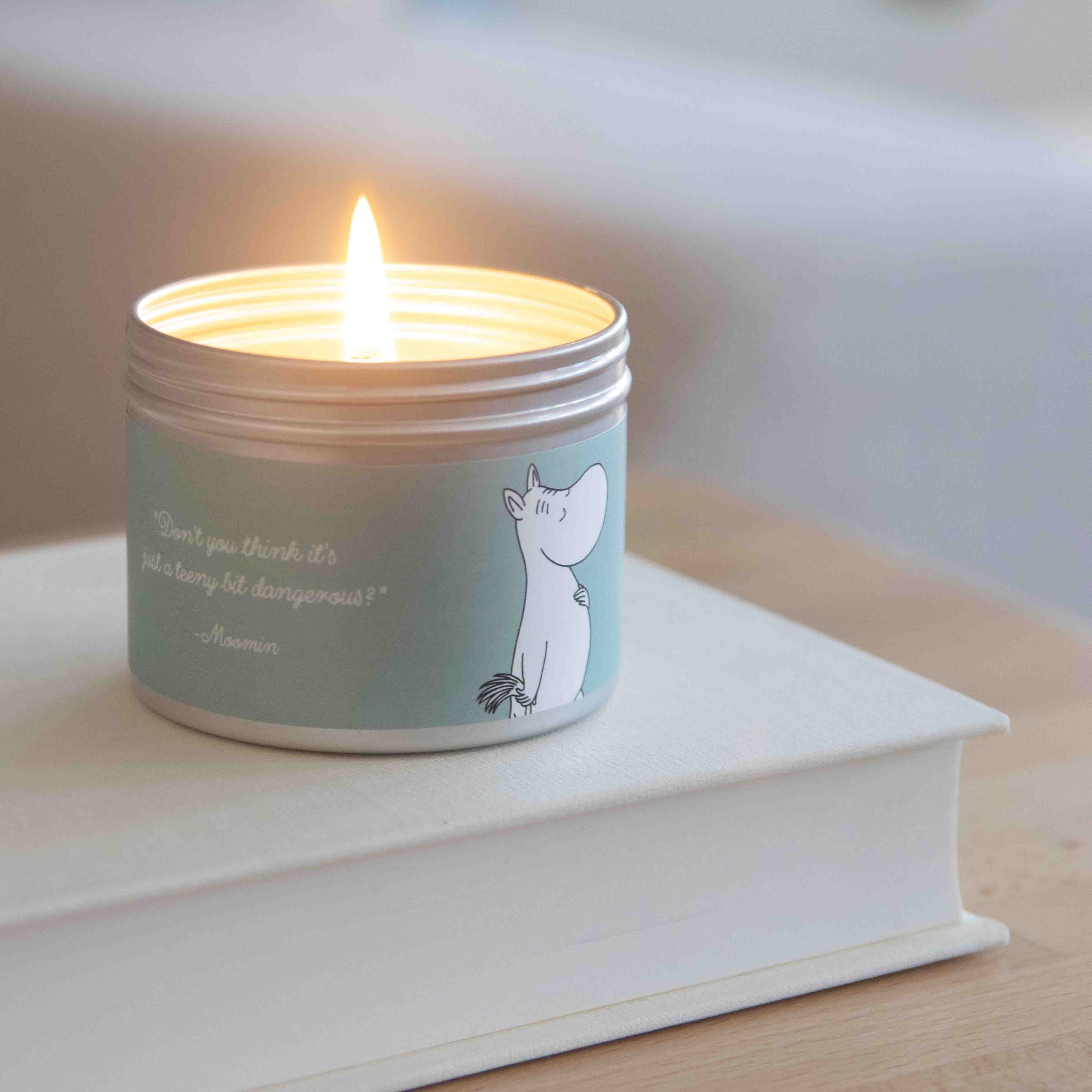 A lit Moomin-themed candle in a silver tin.  The teal label features a Moomin character illustration and quote.