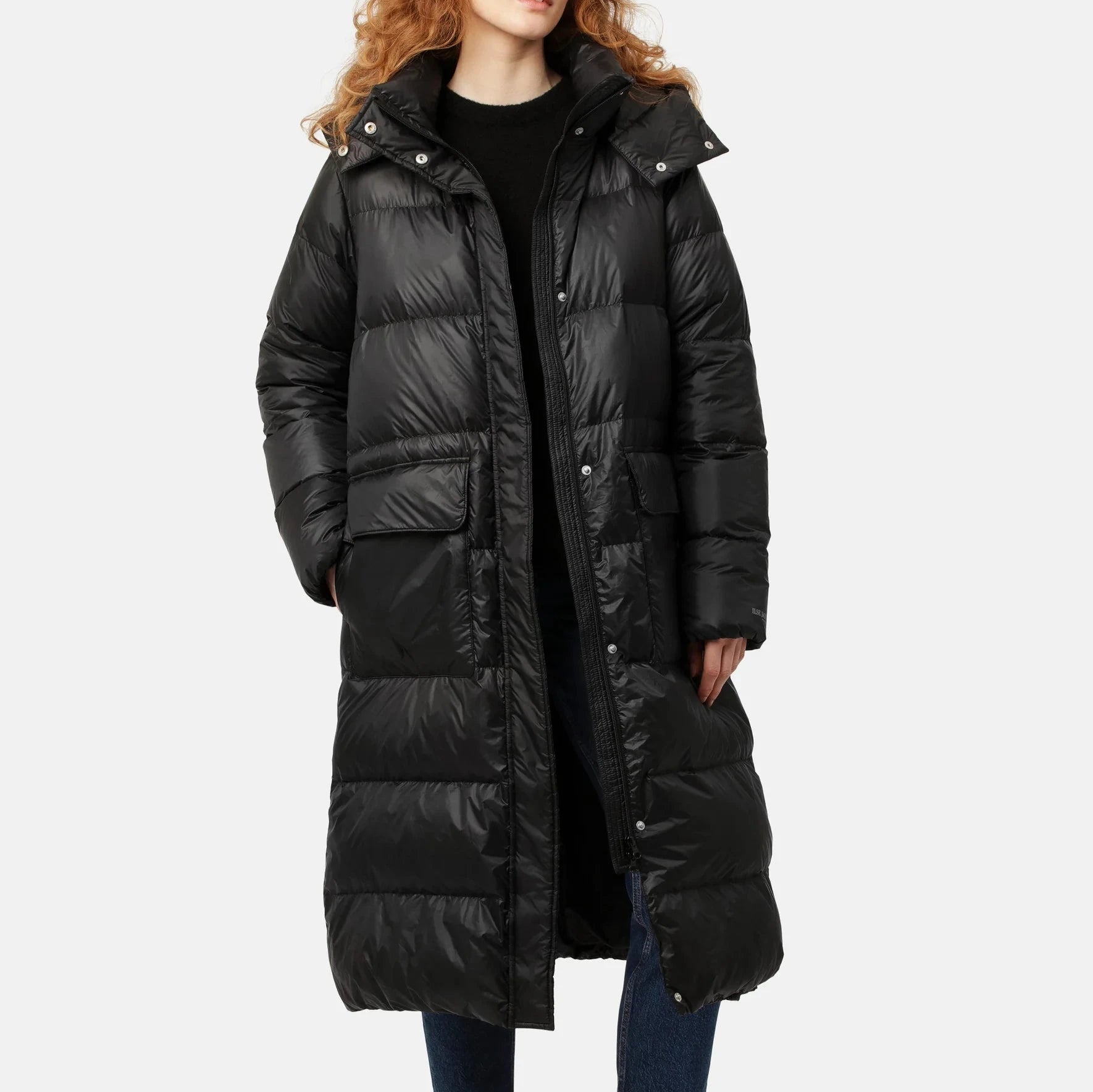 Black long puffer coat with a hood, featuring a shiny, quilted texture and two lower pockets. The model wears jeans underneath.