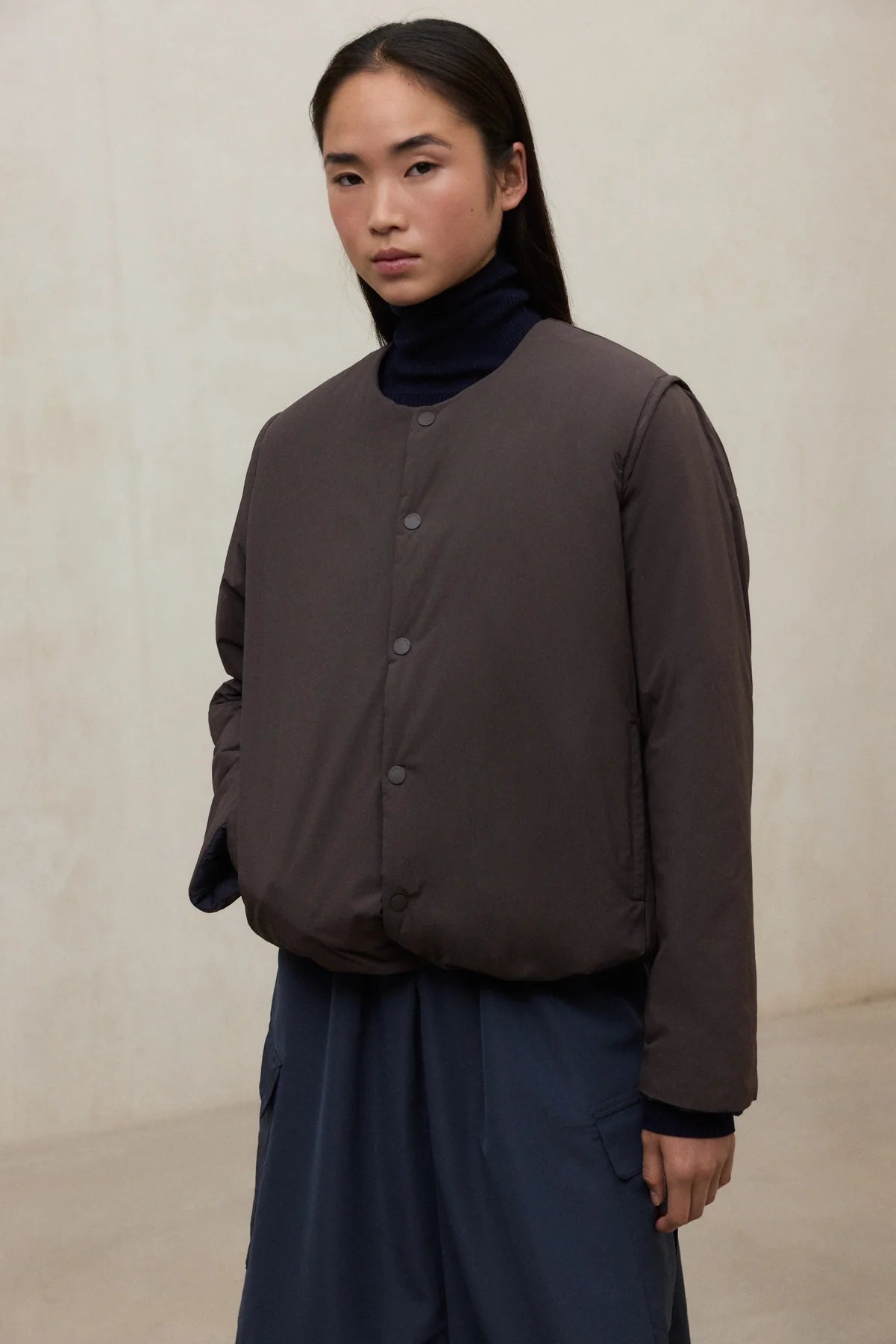 Person wearing a brown jacket over a dark turtleneck and blue pants on a beige background