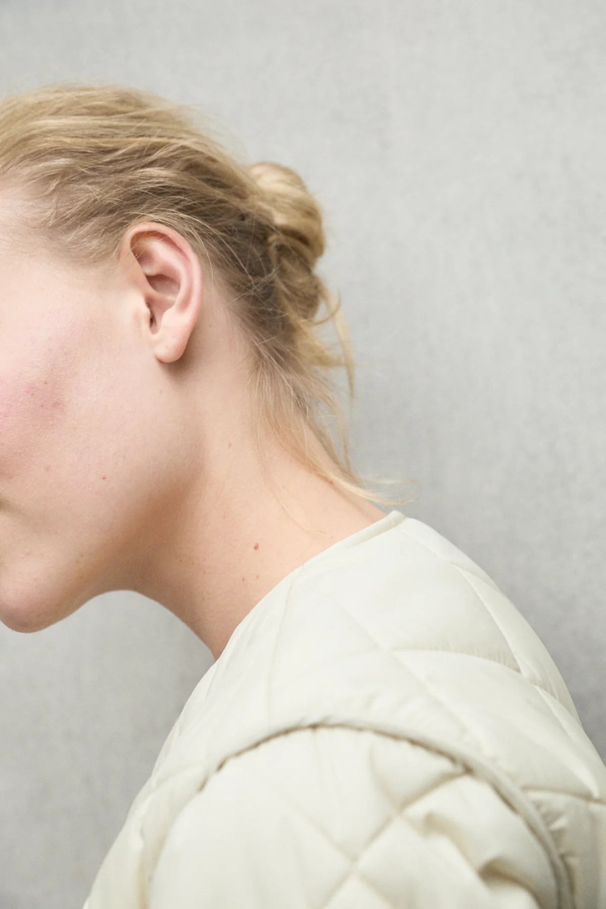 Close-up of a woman's neck and shoulder, wearing a cream-colored quilted jacket. Her blonde hair is styled in a loose bun.
