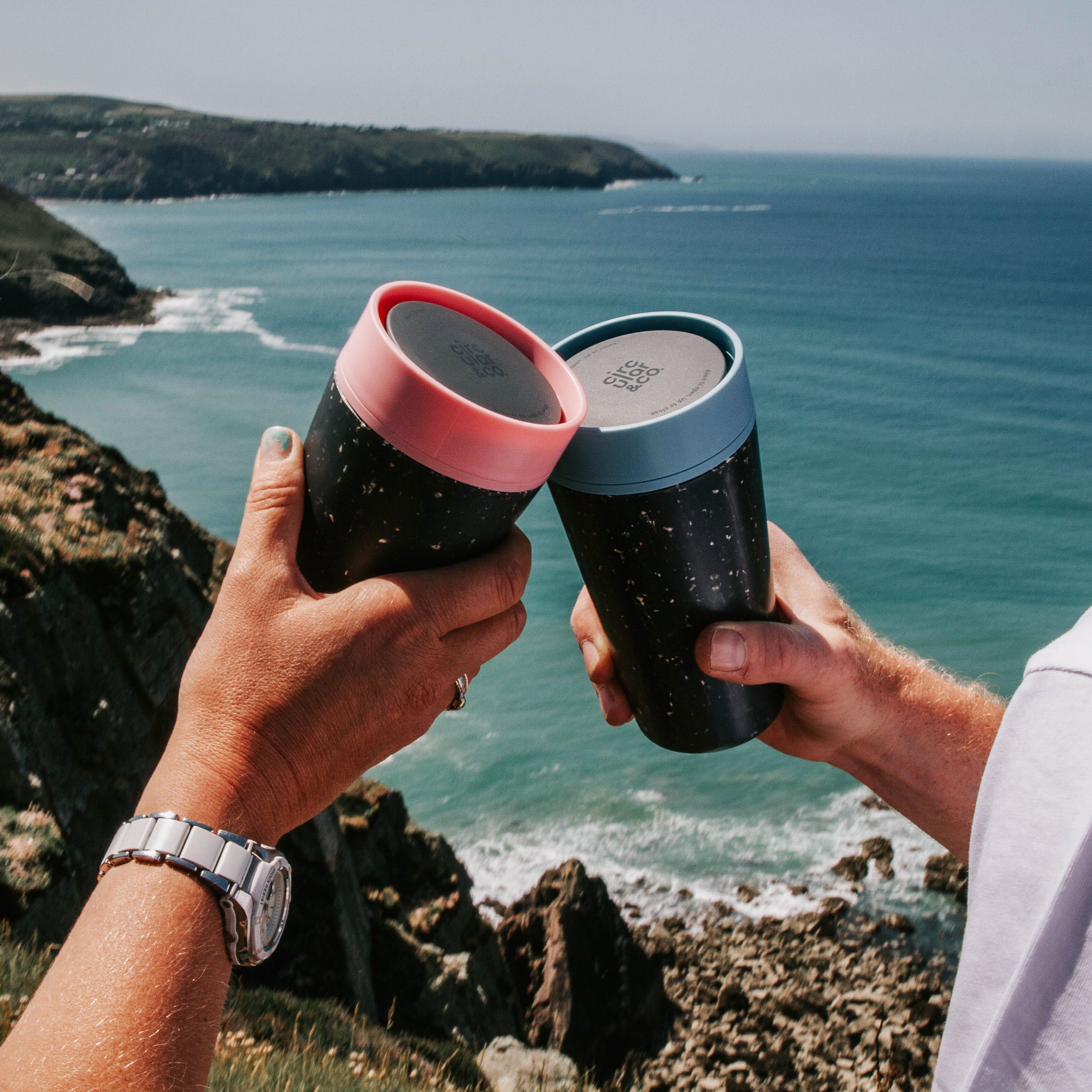 Two black reusable coffee cups with pink and blue lids, held up in a toast, against a blurry ocean backdrop.