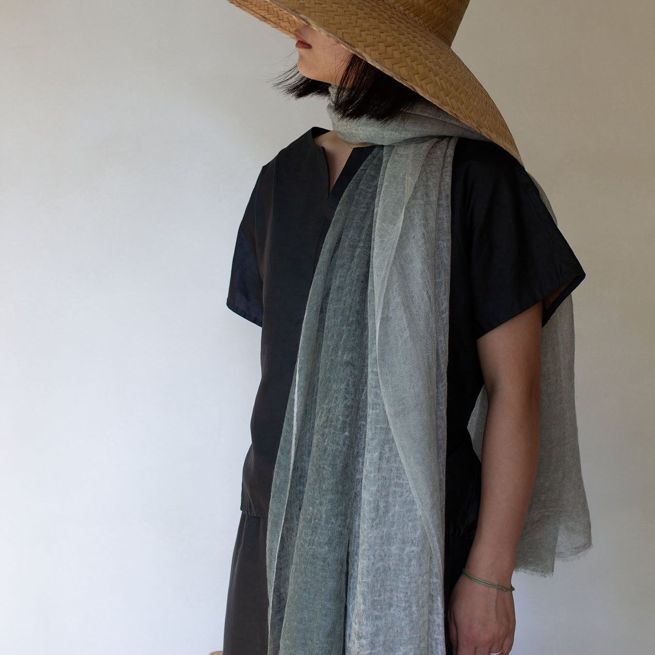 A person wearing a wide-brimmed hat and a macaron ombre cashmere scarf in sage green, paired with a black top.