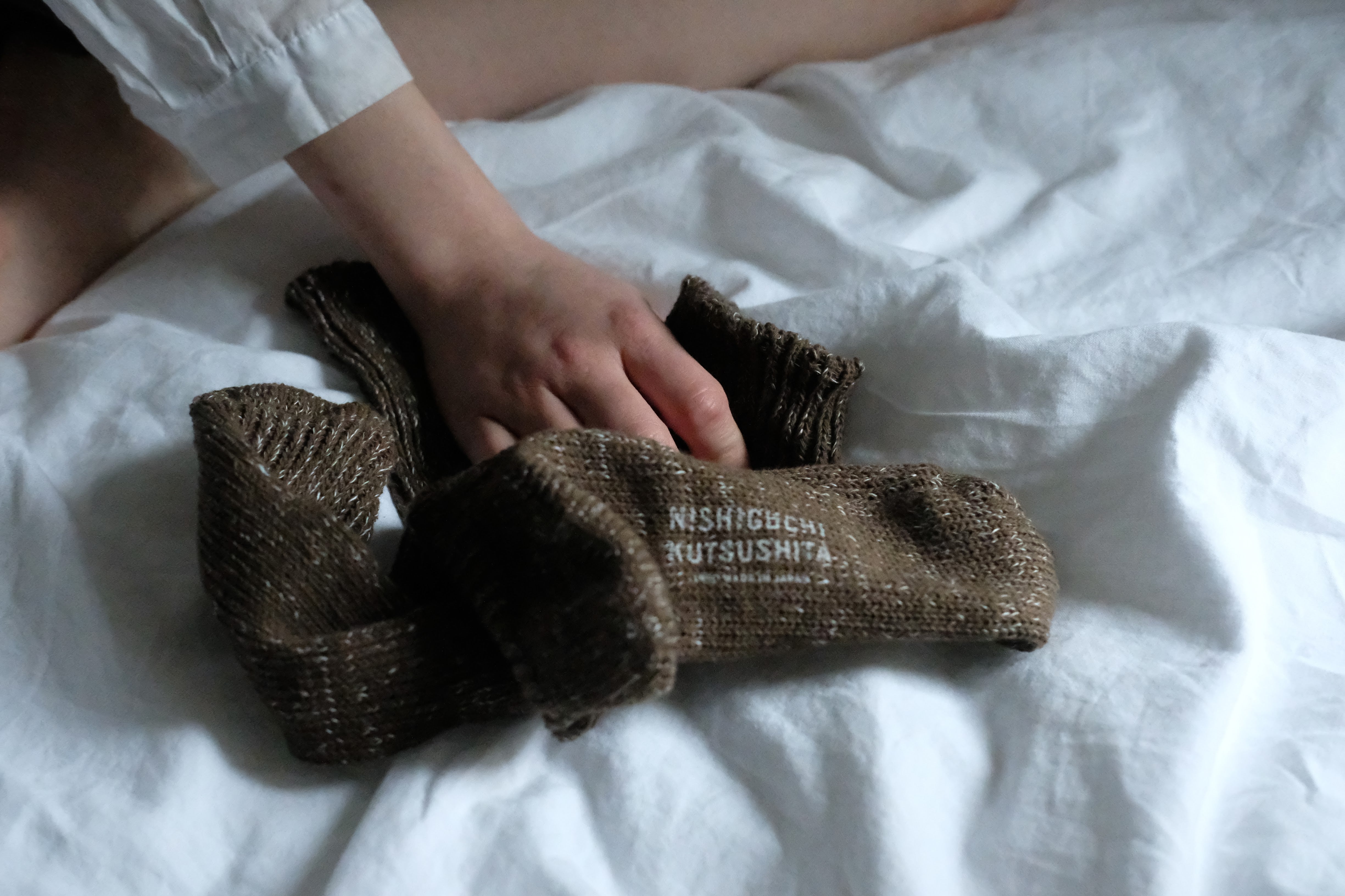 A pair of brown knitted socks with subtle white flecks and "NISHIGUCHI KUTSUSITA" branding lies on a white bedsheet.