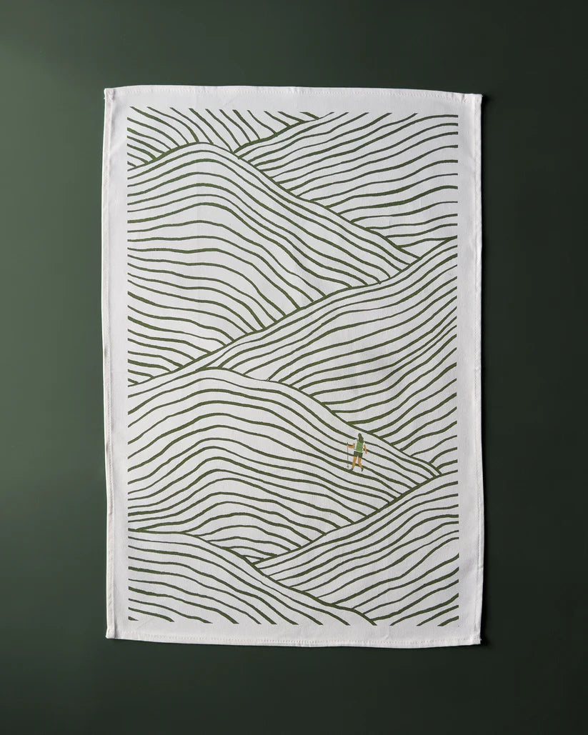 Tea towel with illustration of rolling hills in olive green lines on a white background; a small figure hikes across the