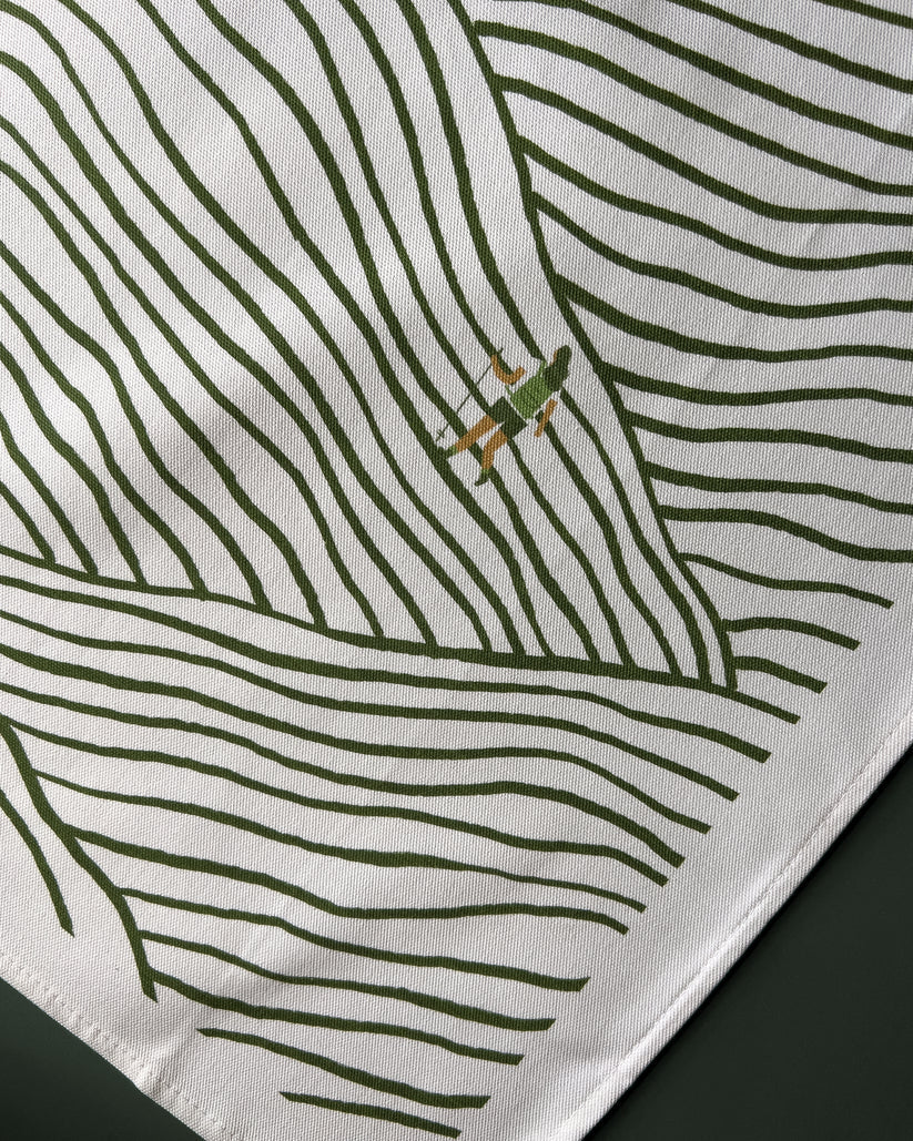 White tea towel with a repeating pattern of olive green lines suggesting hills, and a small illustration of a hiker.