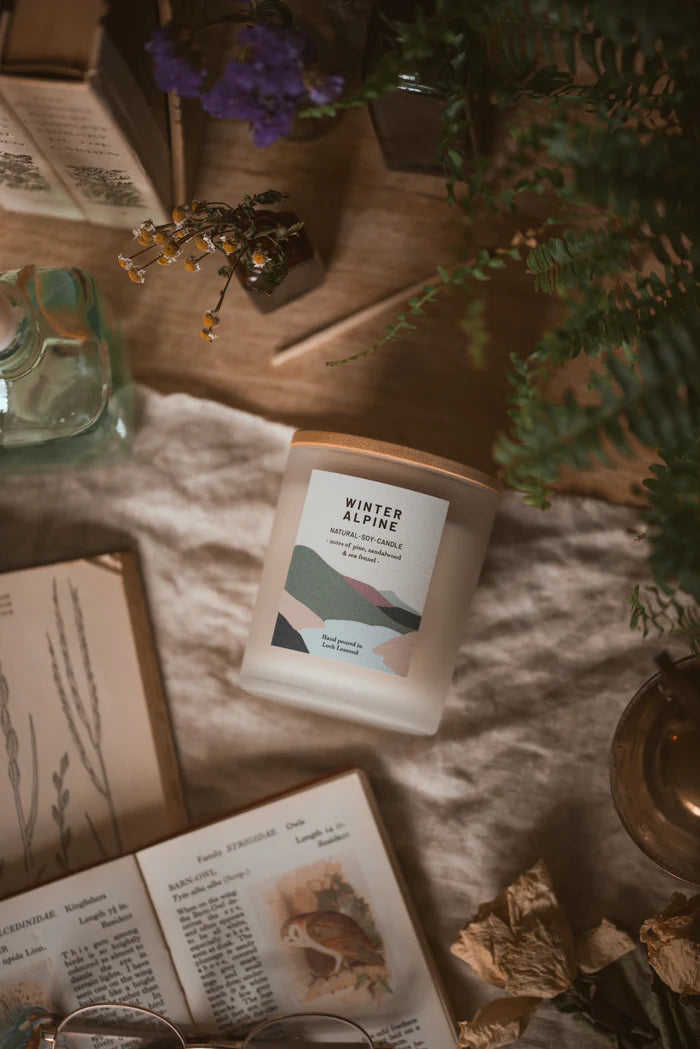 Frosted glass candle with wooden lid; label reads "Winter Alpine," "Natural Soy Candle," and notes of pine, sandalwood, and