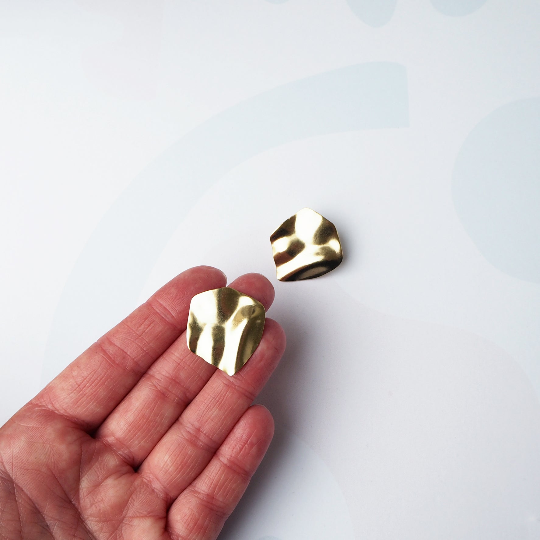 Pair of gold, textured, irregular-shaped earrings.  One earring rests on a hand.